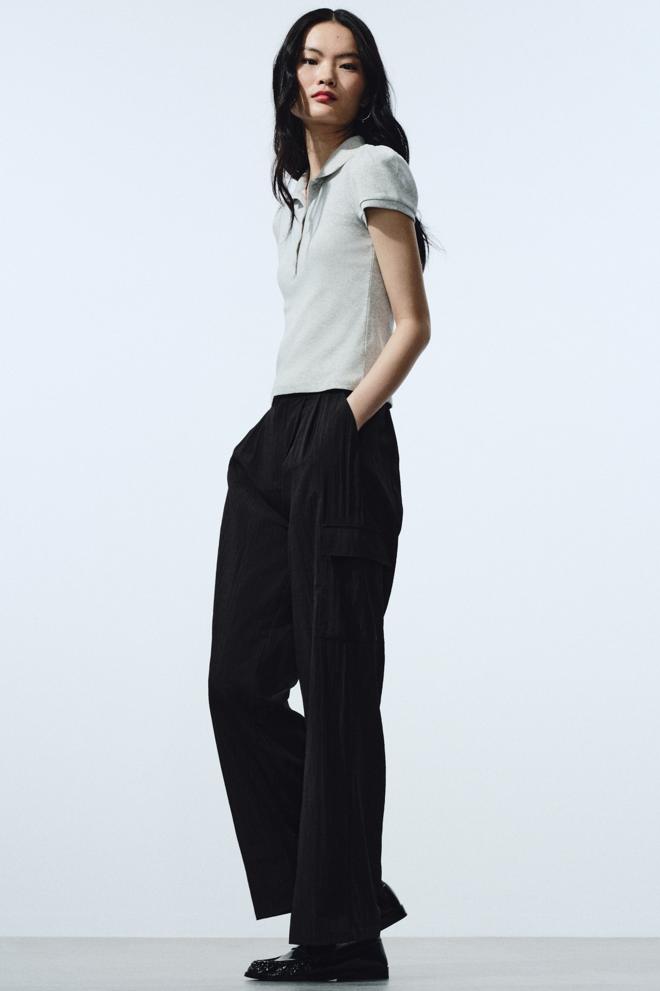 拡大画像を表示: ライトグレーのニットポロシャツは、ギャザー入りのショートスリーブ、クラシックな襟、2つボタンの前立てを備えています。フィット感のある、ややクロップド丈のシルエットです。