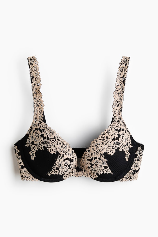 A bra is showcased from the front, highlighting black underwired cups adorned with a beige floral lace overlay. Scalloped lace edges frame the cups and straps, with a small black bow embellishing the centre gore. - 1