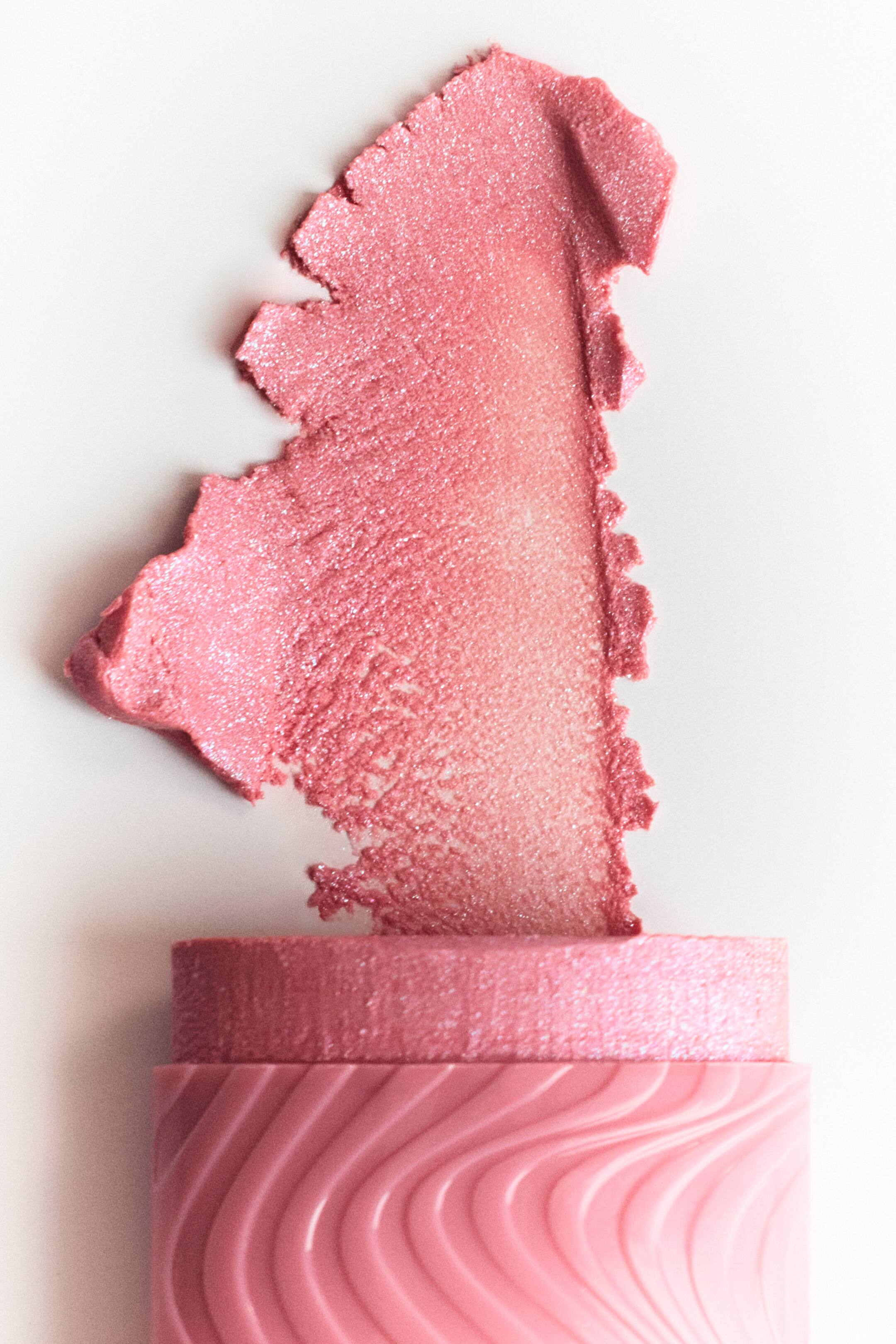 Vis større bilde: En rosa kosmetisk stift viser frem en skimrende, glitrende rosa prøve. Stiftens synlige hylster har et skulpturert bølgemønster.