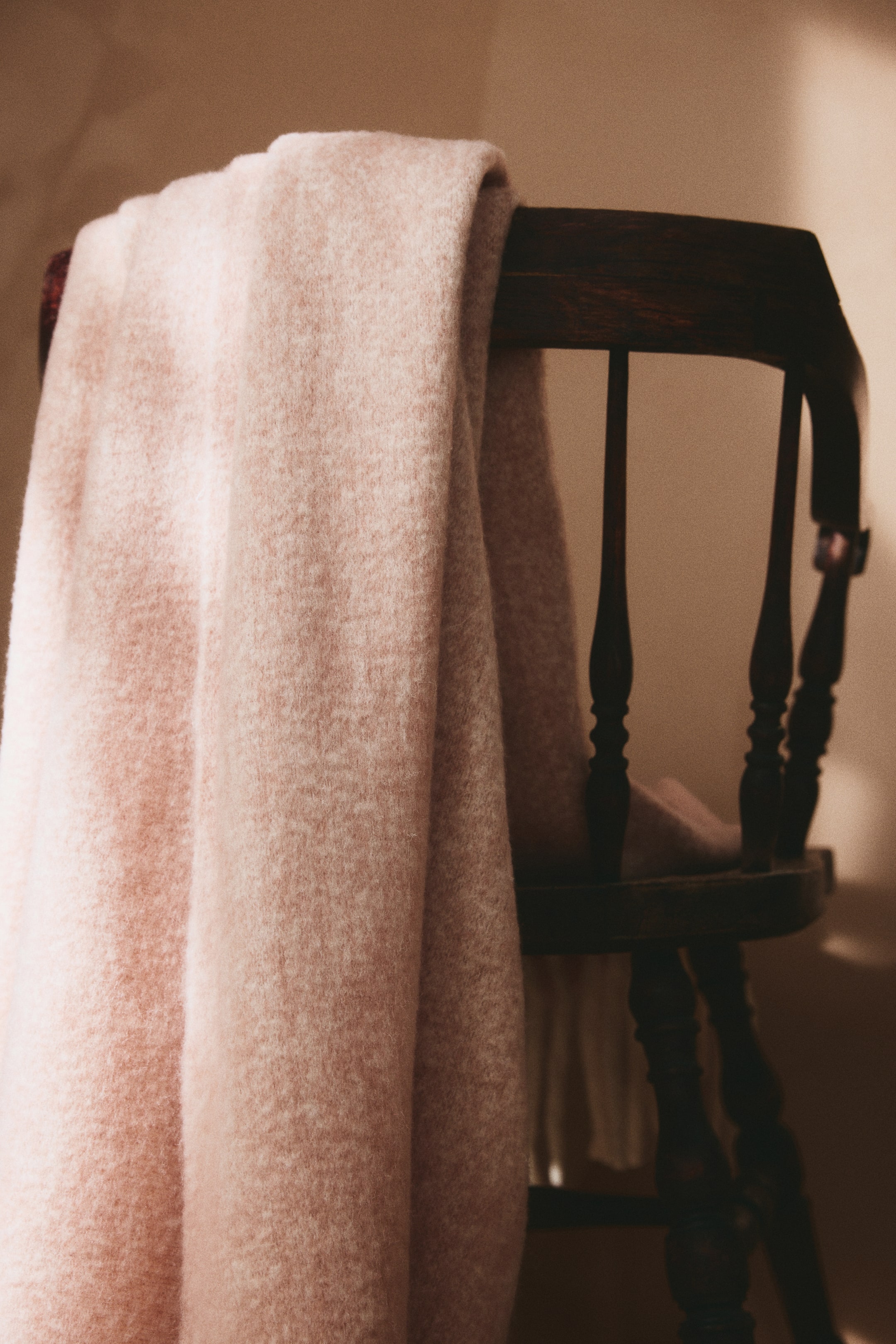 View larger image: A light pink blanket in a soft wool-blend weave with a brushed finish is draped over the back of a dark wooden chair, showcasing its fuzzy, textured surface. Fringes hang from the short side of the folded blanket near the bottom.