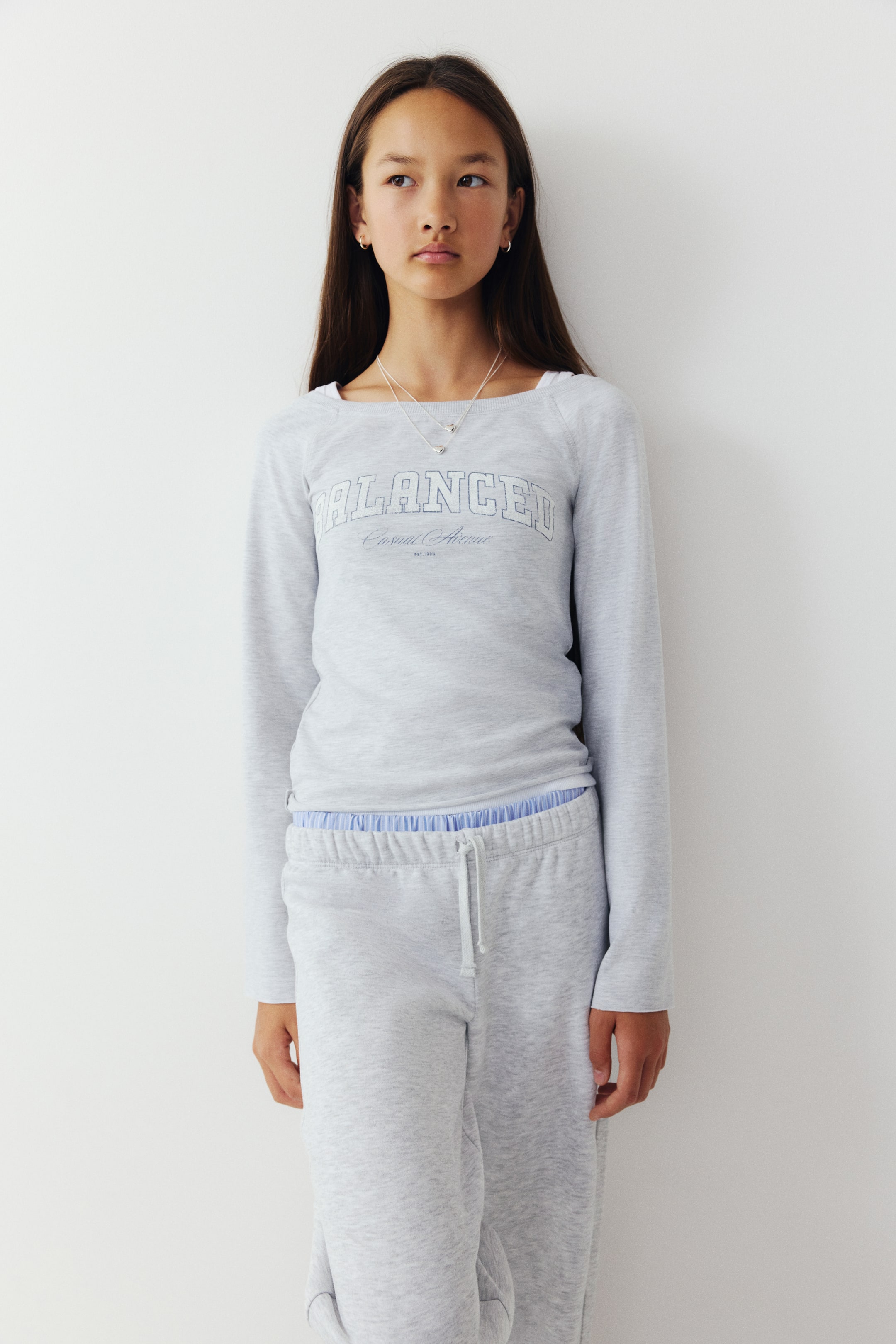 Visa större bild: En ljusgrå sweatshirt i sweattyg visas framifrån och har en ribbad båthals, långa raglanärmar och ett böjt collegeliknande texttryck med ordet "BALANCED" på bröstet. Tröjan stylas med ett lager av en vit topp undertill och matchande ljusgrå sweatpants.