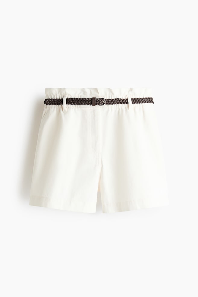I pantaloncini bianchi presentano una vita arricciata e tasche laterali visibili, mostrati frontalmente. Una cintura intrecciata marrone scuro con fibbia in metallo è infilata nei passanti della cintura. - 1