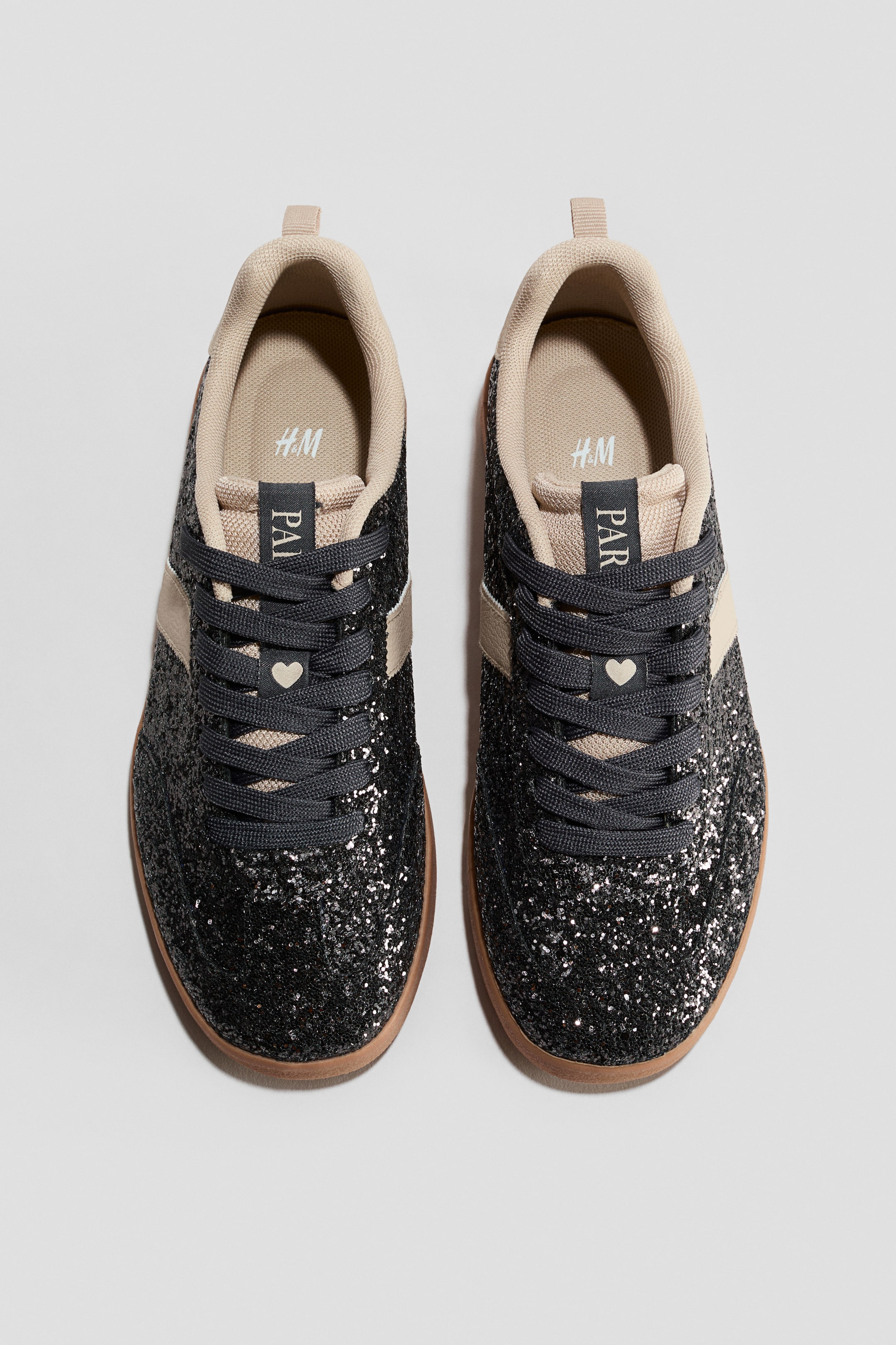 Agrandir l'image: Une paire de baskets avec des dessus en paillettes noires et des bandes latérales beige clair, des lacets gris foncé et un petit détail en forme de cœur beige. Les baskets affichent une doublure intérieure beige avec un logo H&M, une languette marquée "PAR", et des semelles de couleur gomme brune.