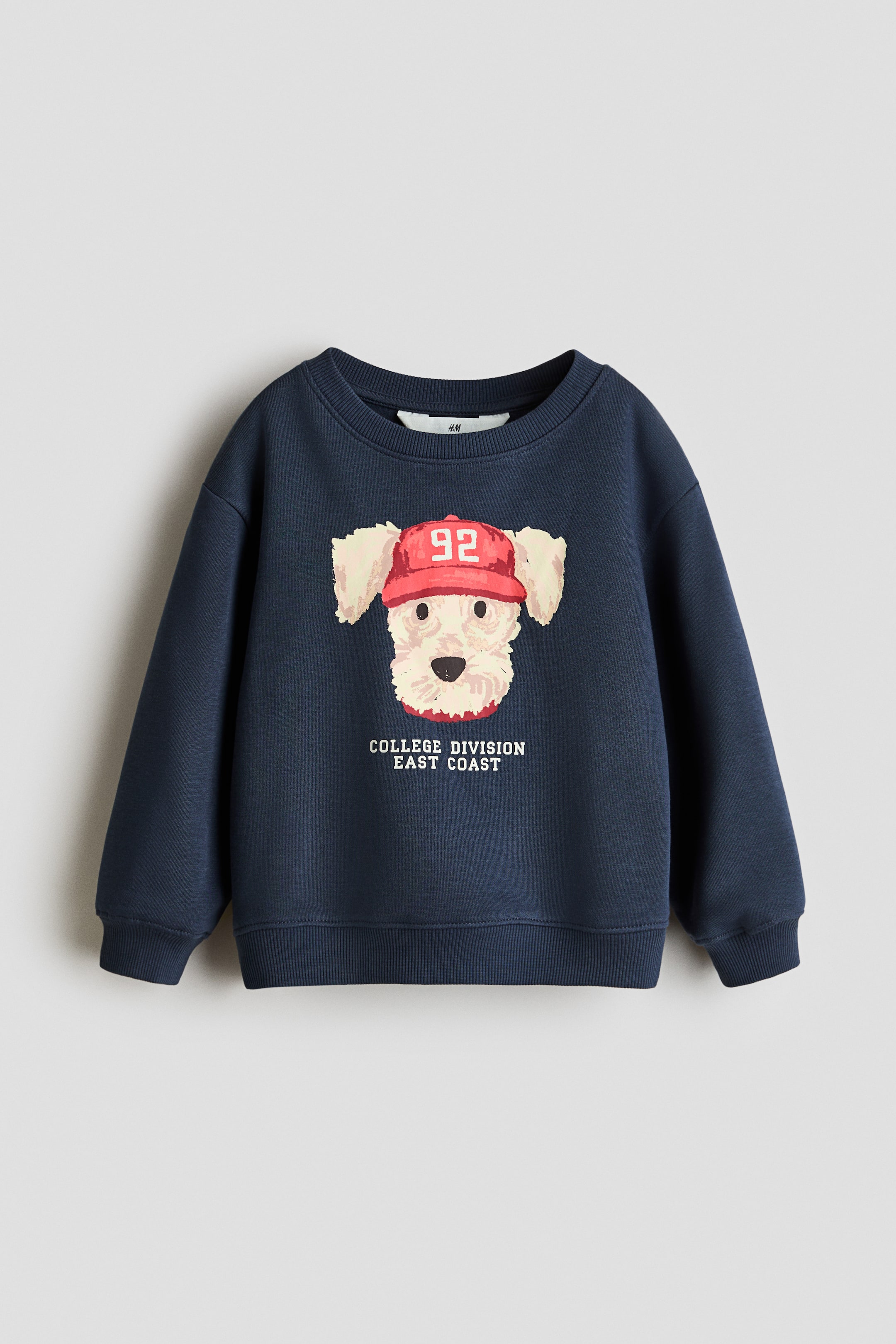 View larger image: A navy blue sweatshirt features a graphic print of a white dog's head wearing a red "92" cap, with "COLLEGE DIVISION EAST COAST" text below on the front. It has a round, rib-trimmed neckline, dropped shoulders, long sleeves, and ribbing at the cuffs and hem.