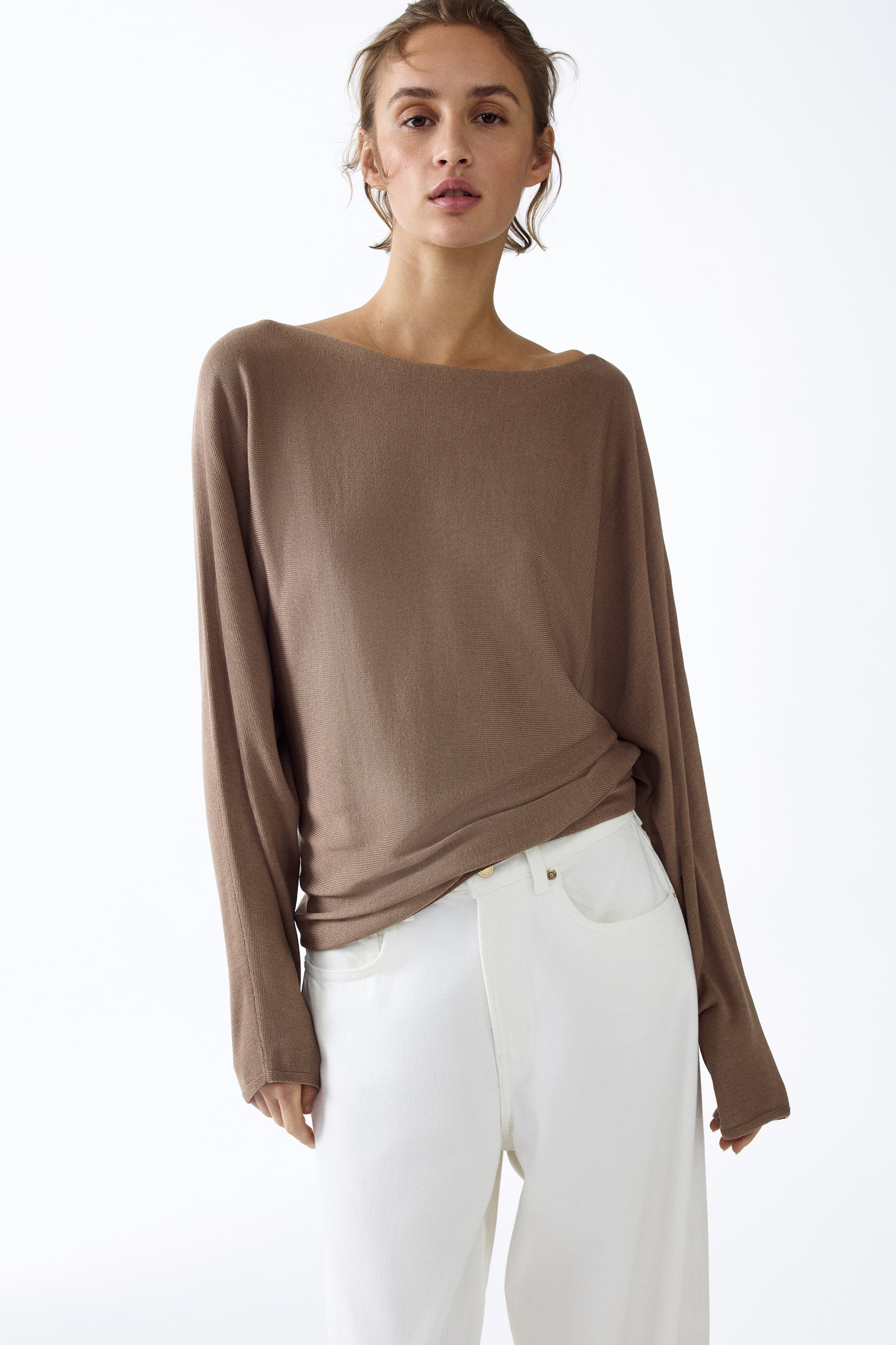 View larger image: A brown long-sleeved top is showcased, featuring a wide boat neck and a relaxed, draped fit with tapering sleeves and simple cuffs. The top is styled loosely over white trousers, with its hem partially tucked on the left side.