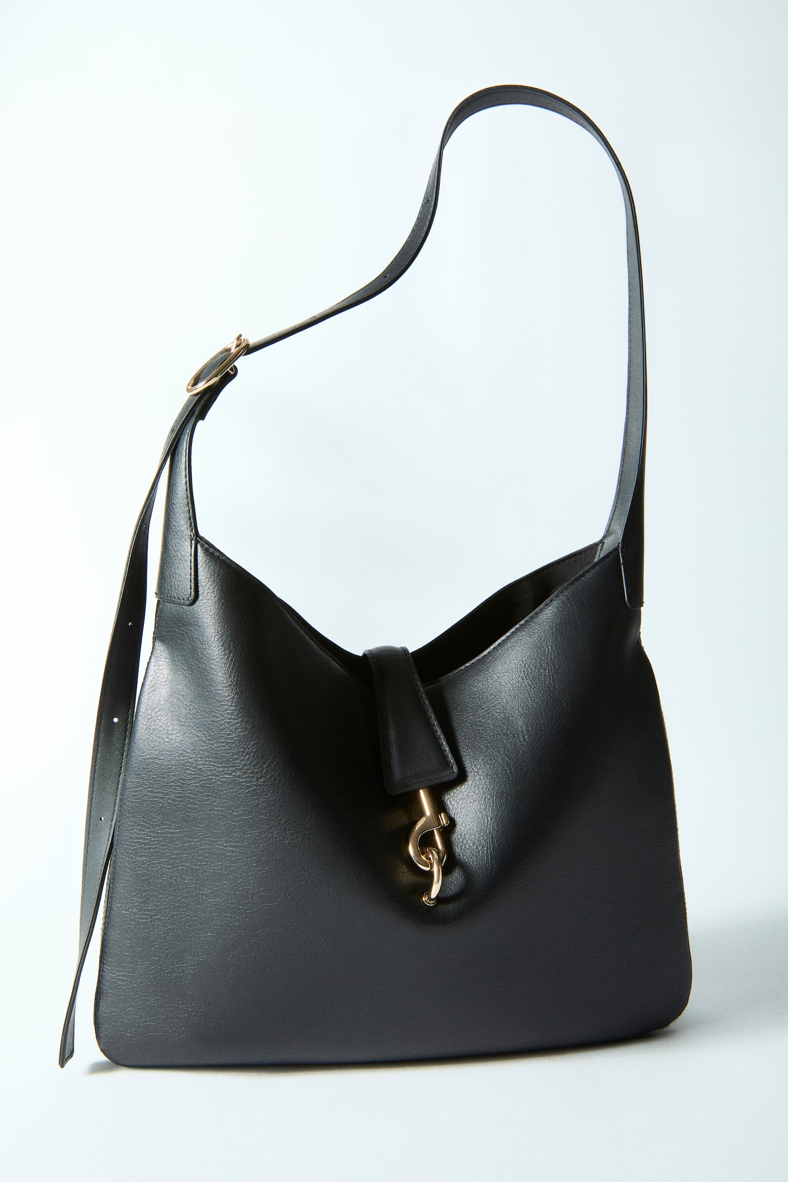 A black handbag is displayed front-facing, boasting a single shoulder strap with a gold-toned buckle. It highlights a grained texture and a flap closure secured by a gold-toned dog clip. - 1