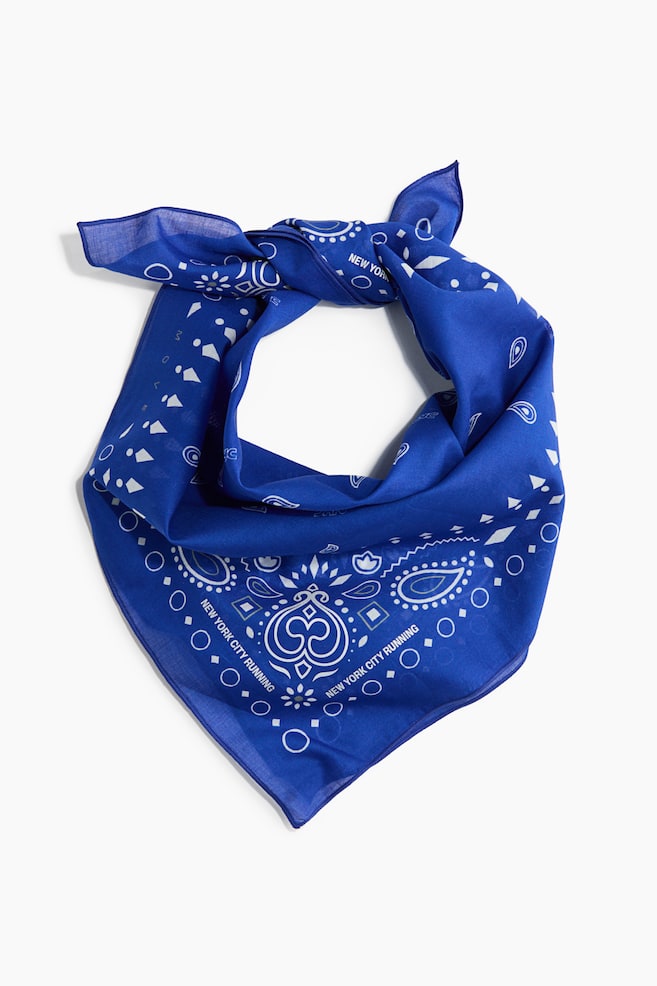 A royal blue bandana is presenting a front view, styled in a circular loop with the pointed corner hanging downward. The lightweight fabric is showcasing white printed designs, including paisley motifs, geometric shapes, and the text "NEW YORK CITY RUNNING" along the lower borders. - 1
