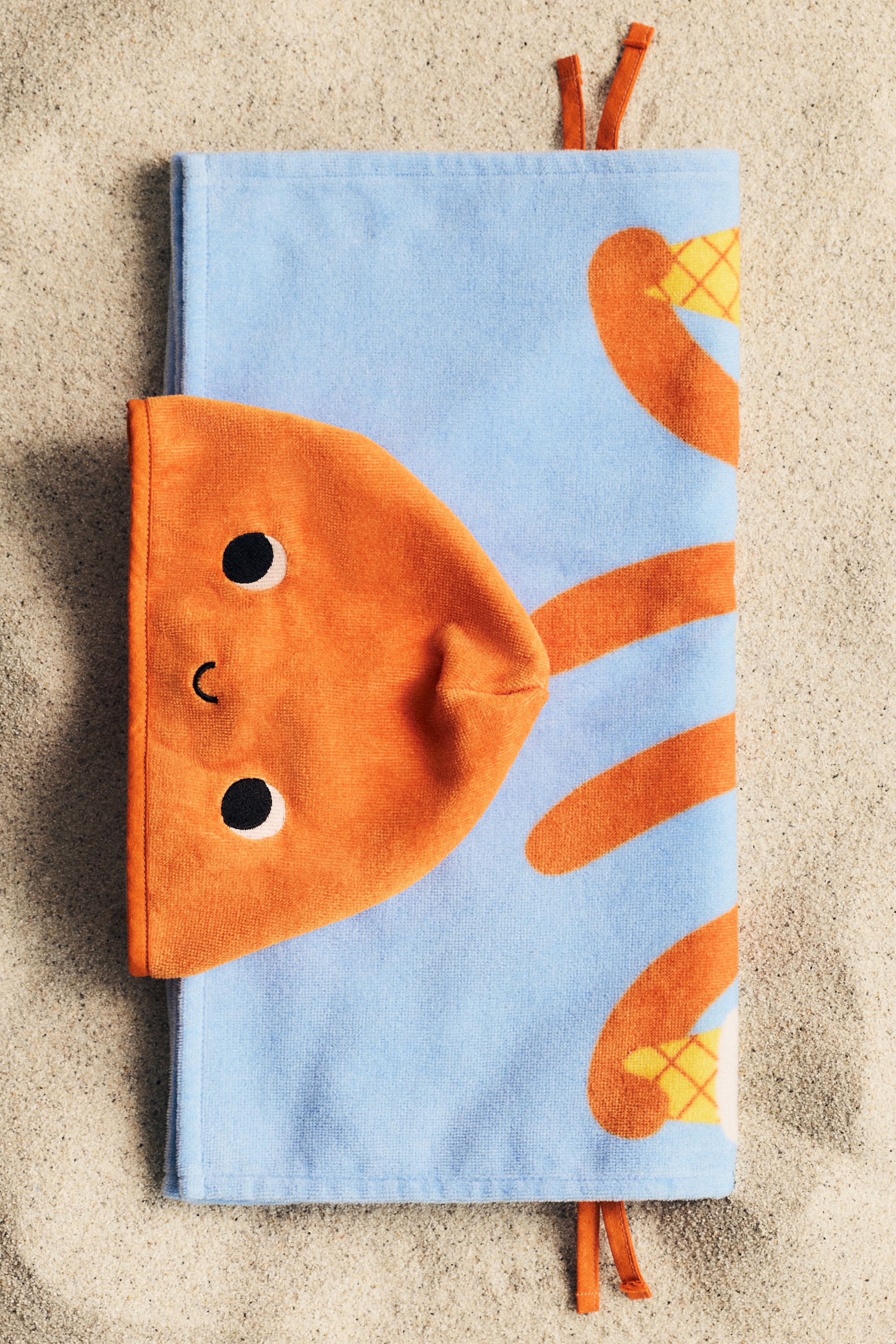 View larger image: A light blue and orange animal towel cape is shown from the front, featuring an orange velour hood with embroidered black eyes and a small black mouth. The light blue towel body is adorned with orange curved shapes and yellow checkered details, with orange narrow ties extending from the top and bottom edges.