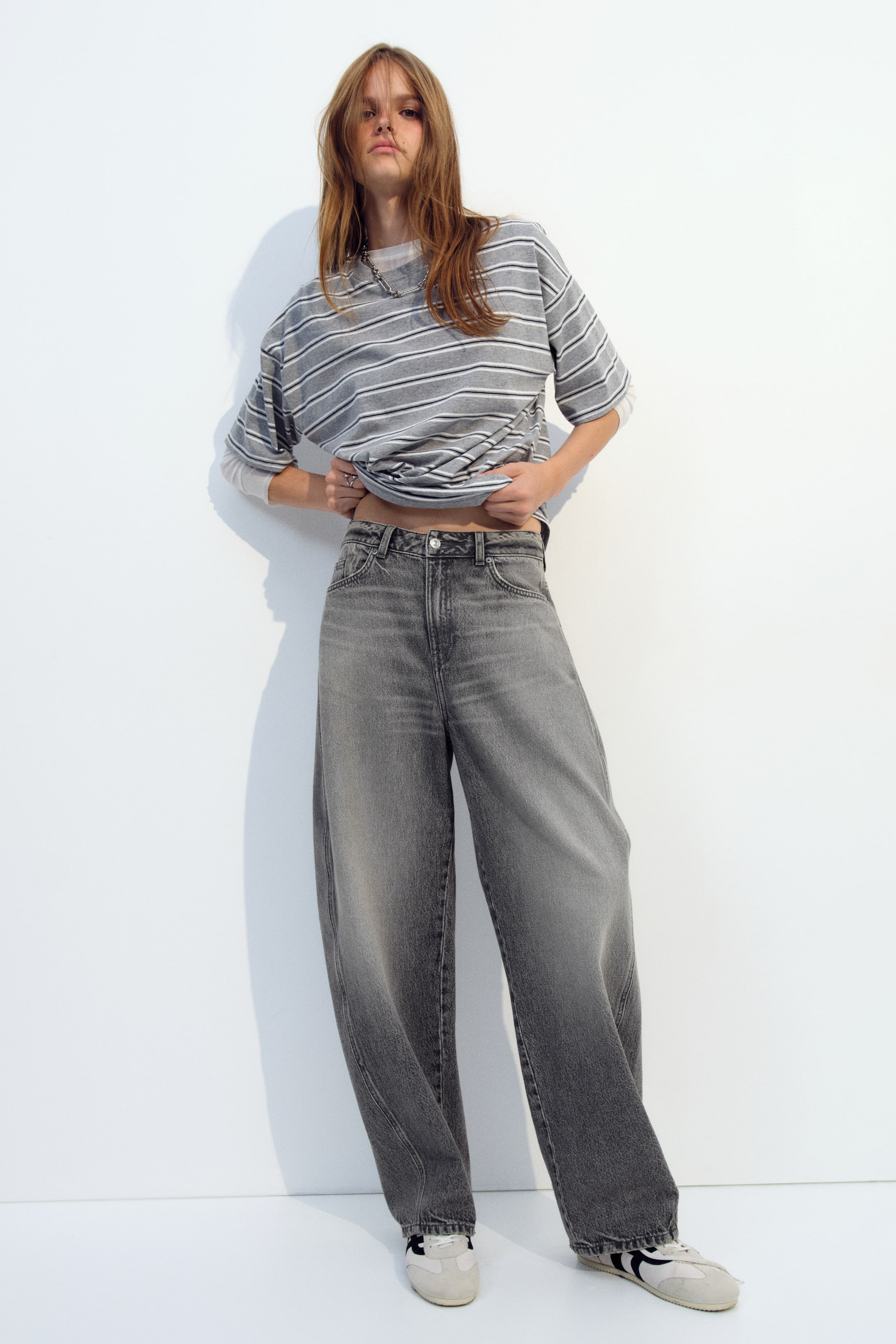 Agrandir l'image: Un jean en denim gris délavé à taille haute, doté d’une jambe courbe et volumineuse, est porté par le mannequin, qui relève l’ourlet d’un t-shirt rayé pour mettre en valeur la ceinture boutonnée. Le jean ample tombe jusqu’aux pieds et est associé à des baskets blanches.