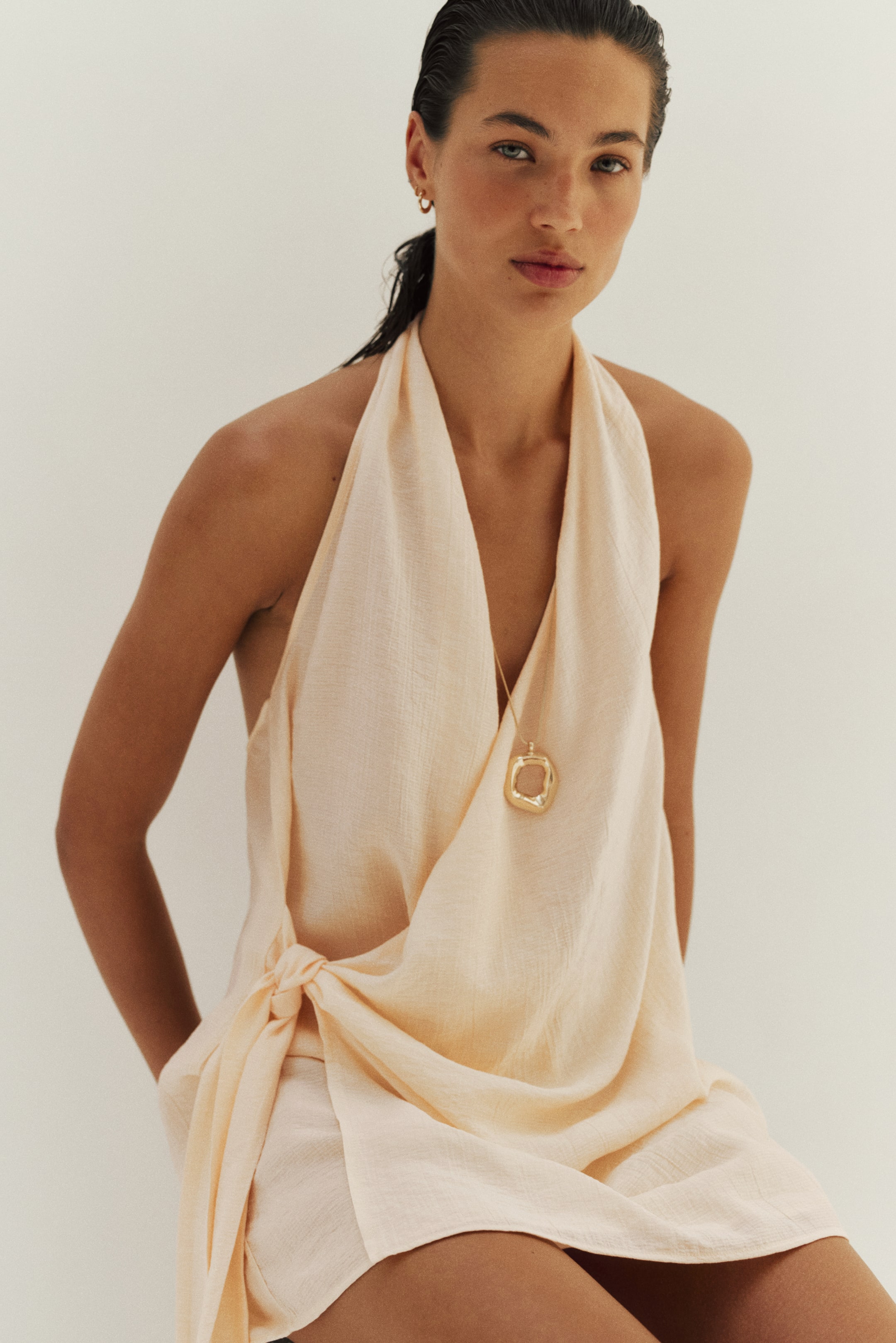 View larger image: A light peach textured halter top features a draped front and a knot detail at the left waist, paired with matching shorts visible underneath. A gold-toned irregular pendant necklace rests on the top.