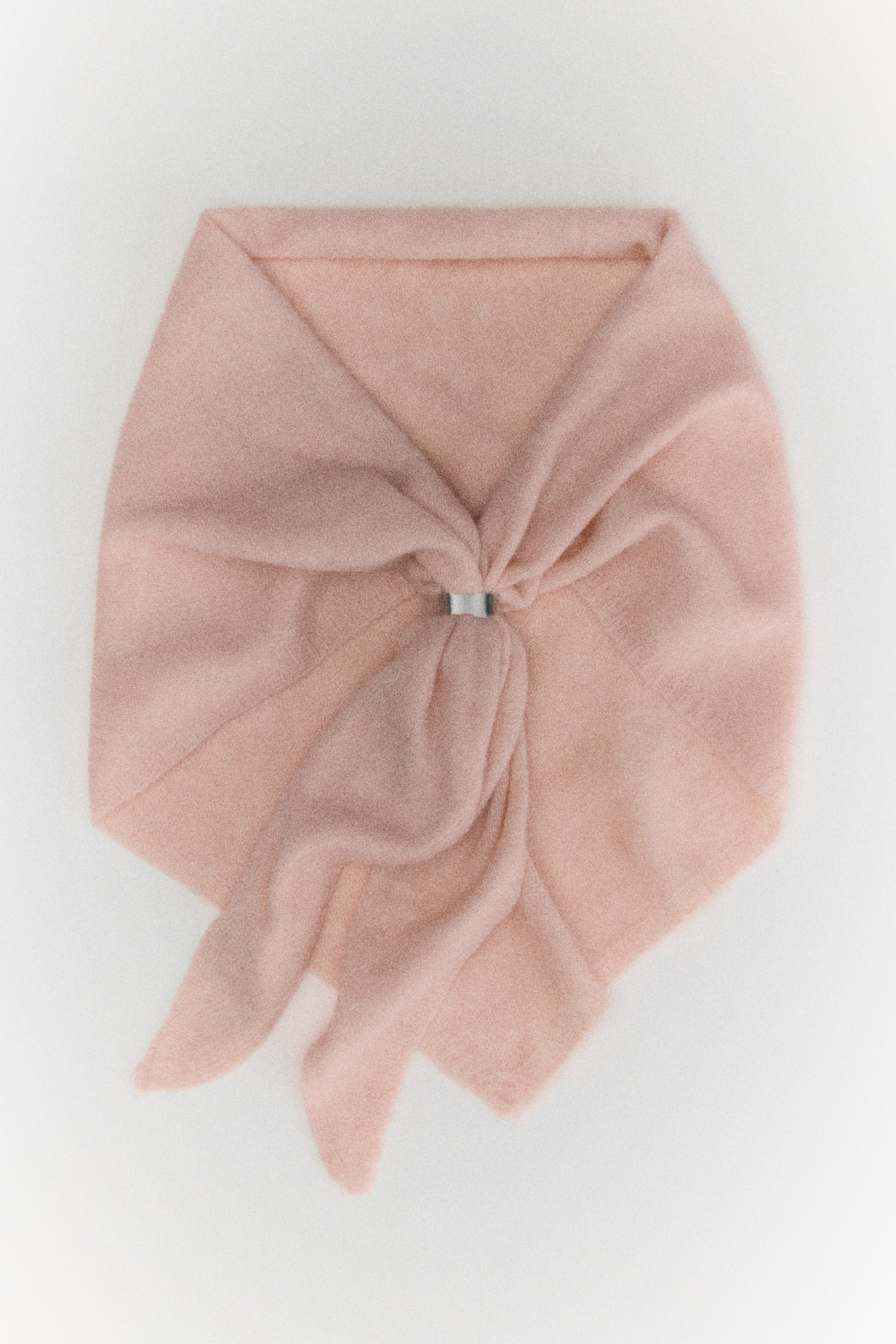 View larger image: A large triangular scarf in a soft, light pink knit is shown from the front, featuring a silver-tone metal ring cinching the fabric together at the centre. The draped ends cascade downwards, showcasing the narrow, ribbed trim along the edges.