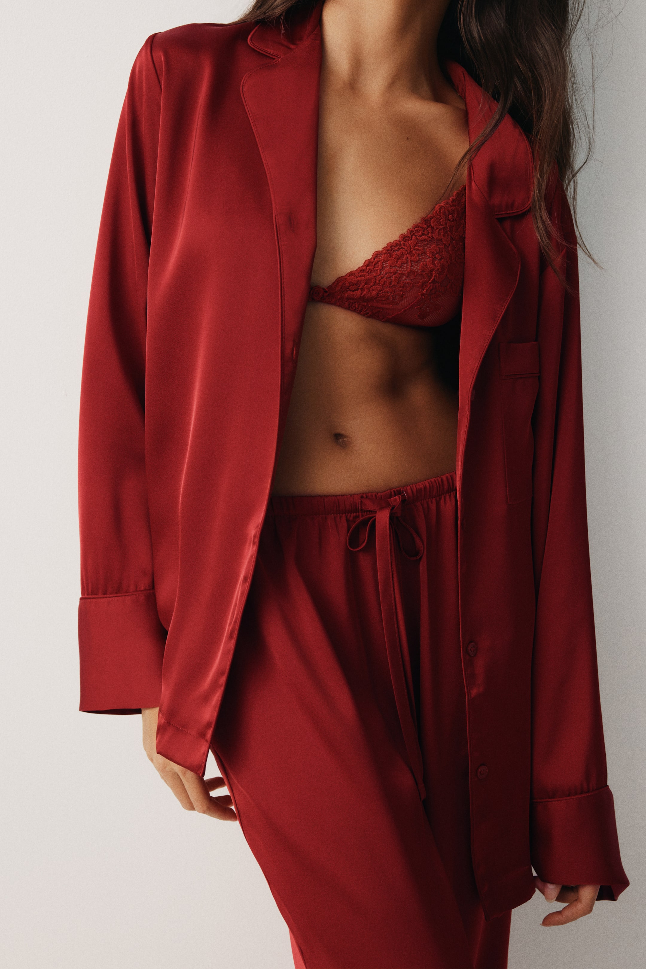 View larger image: A red satin pajama shirt is opening to reveal a matching red lace triangle bralette and red drawstring pajama pants. The long-sleeved shirt is featuring wide cuffs, and the pants are showing a tie-front drawstring waist.
