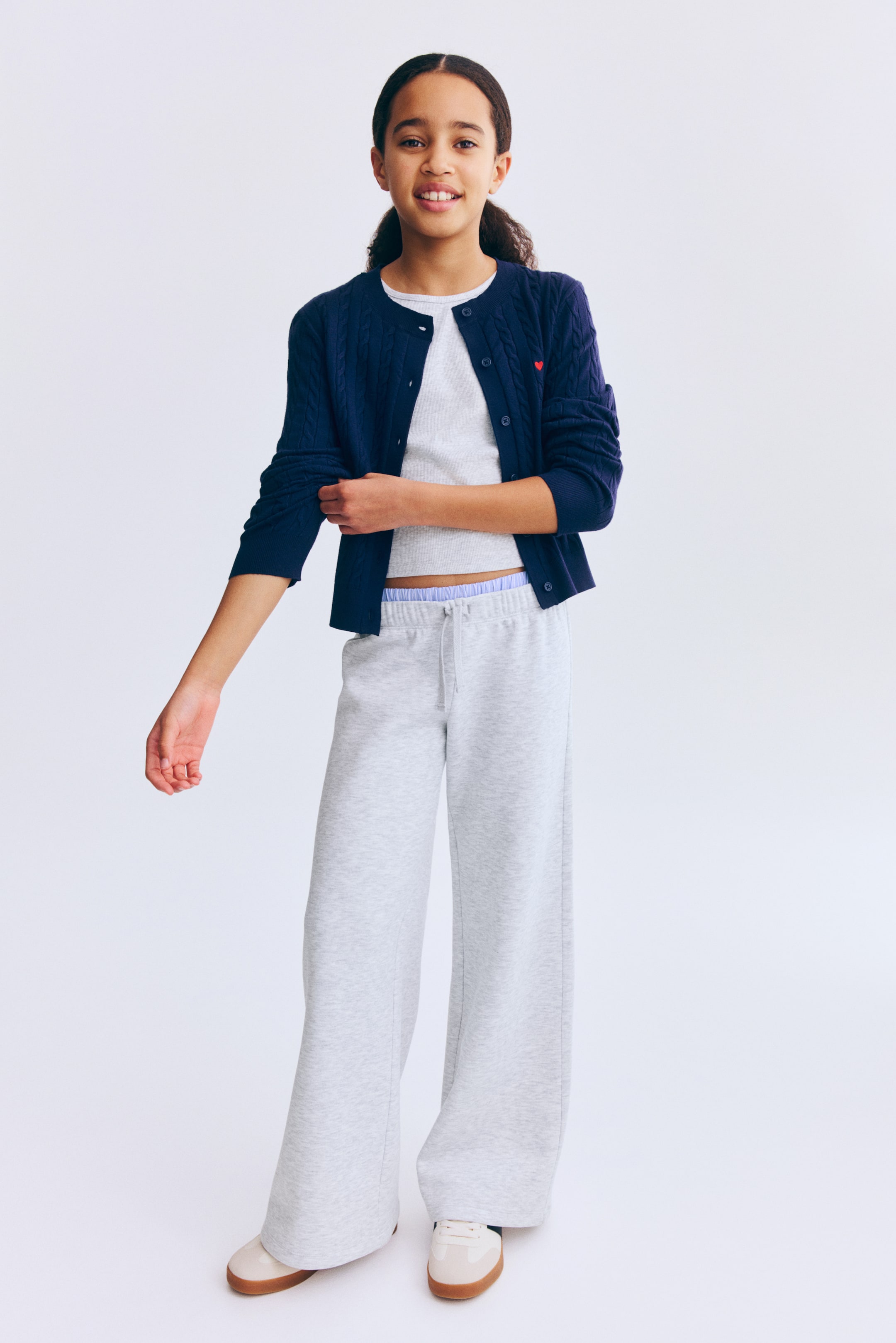 Agrandir l'image: Un cardigan bleu marine à torsades, présenté de face, présente une encolure ronde, des poignets côtelés et un ourlet côtelé. Il est doté d’un boutonnage frontal et d’une petite broderie en forme de cœur rouge sur la poitrine gauche, et est porté ouvert par-dessus un haut gris clair.