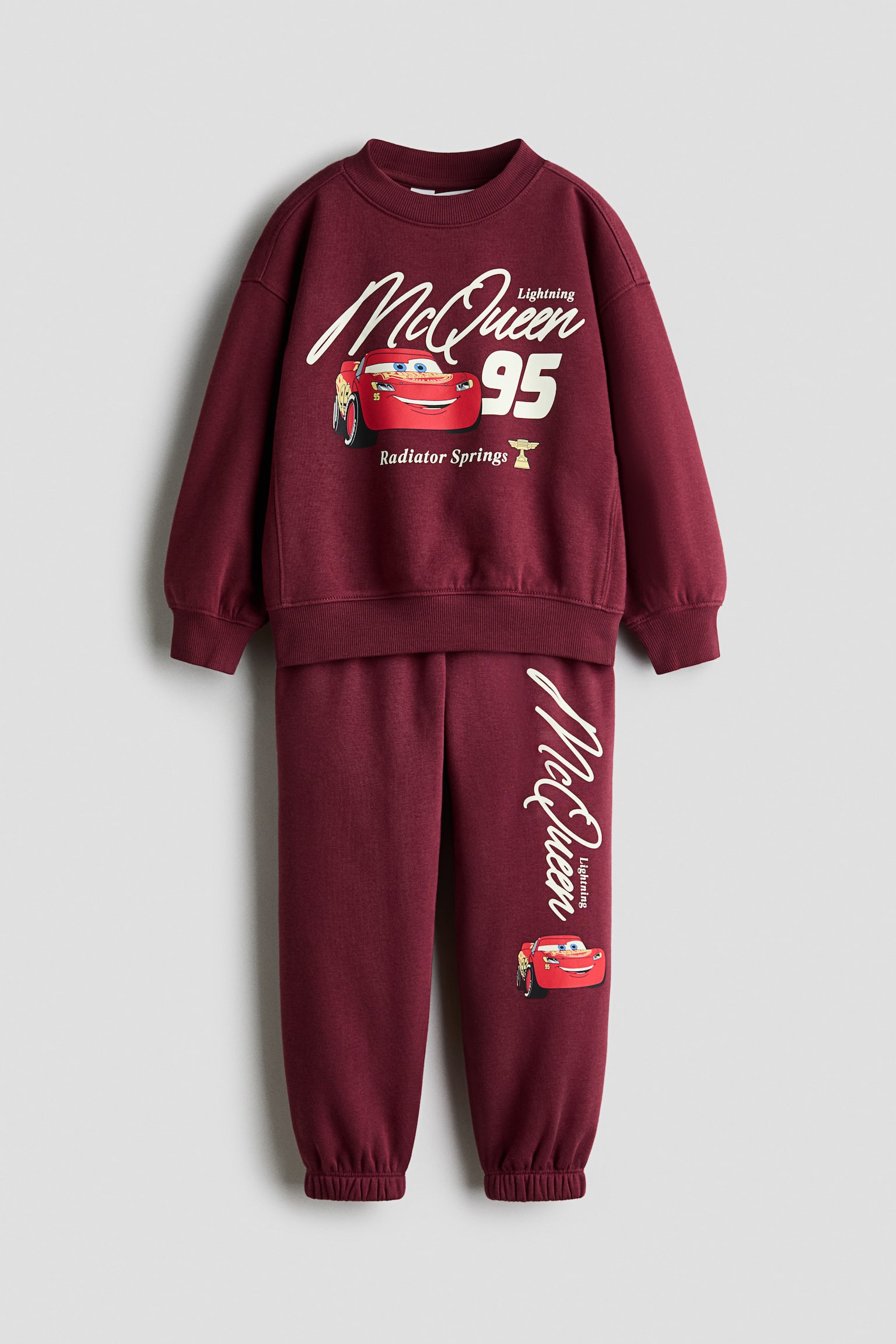 A front view is showcasing a burgundy sweat fabric sweater and matching track pants set featuring graphic prints of Lightning McQueen. The long-sleeve sweater is displaying a ribbed crew neckline, dropped shoulders, and ribbed cuffs and hem, while the coordinating track pants are featuring an elasticated waistband and elasticated ankle cuffs.