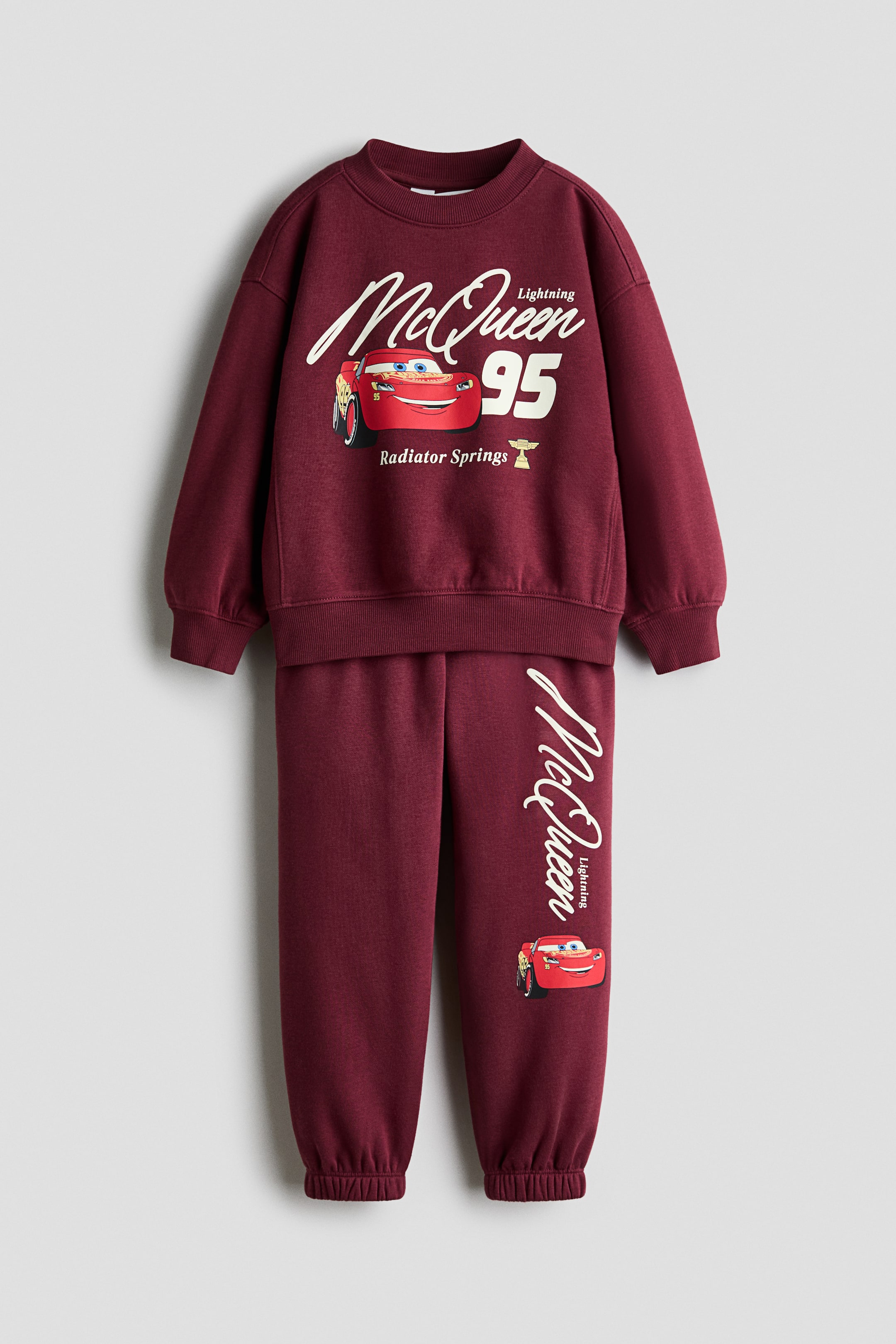 View larger image: A front view showcases a burgundy sweatshirt set made of sweat fabric, featuring graphic prints of Lightning McQueen. The long-sleeve sweatshirt displays a ribbed crew neckline, dropped shoulders, and ribbed cuffs and hem, while the coordinating track pants feature an elasticated waistband and elasticated ankle cuffs.