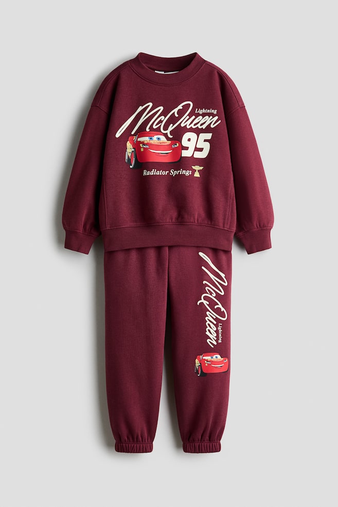 A front view is showcasing a burgundy sweat fabric sweater and matching track pants set featuring graphic prints of Lightning McQueen. The long-sleeve sweater is displaying a ribbed crew neckline, dropped shoulders, and ribbed cuffs and hem, while the coordinating track pants are featuring an elasticated waistband and elasticated ankle cuffs. - 1