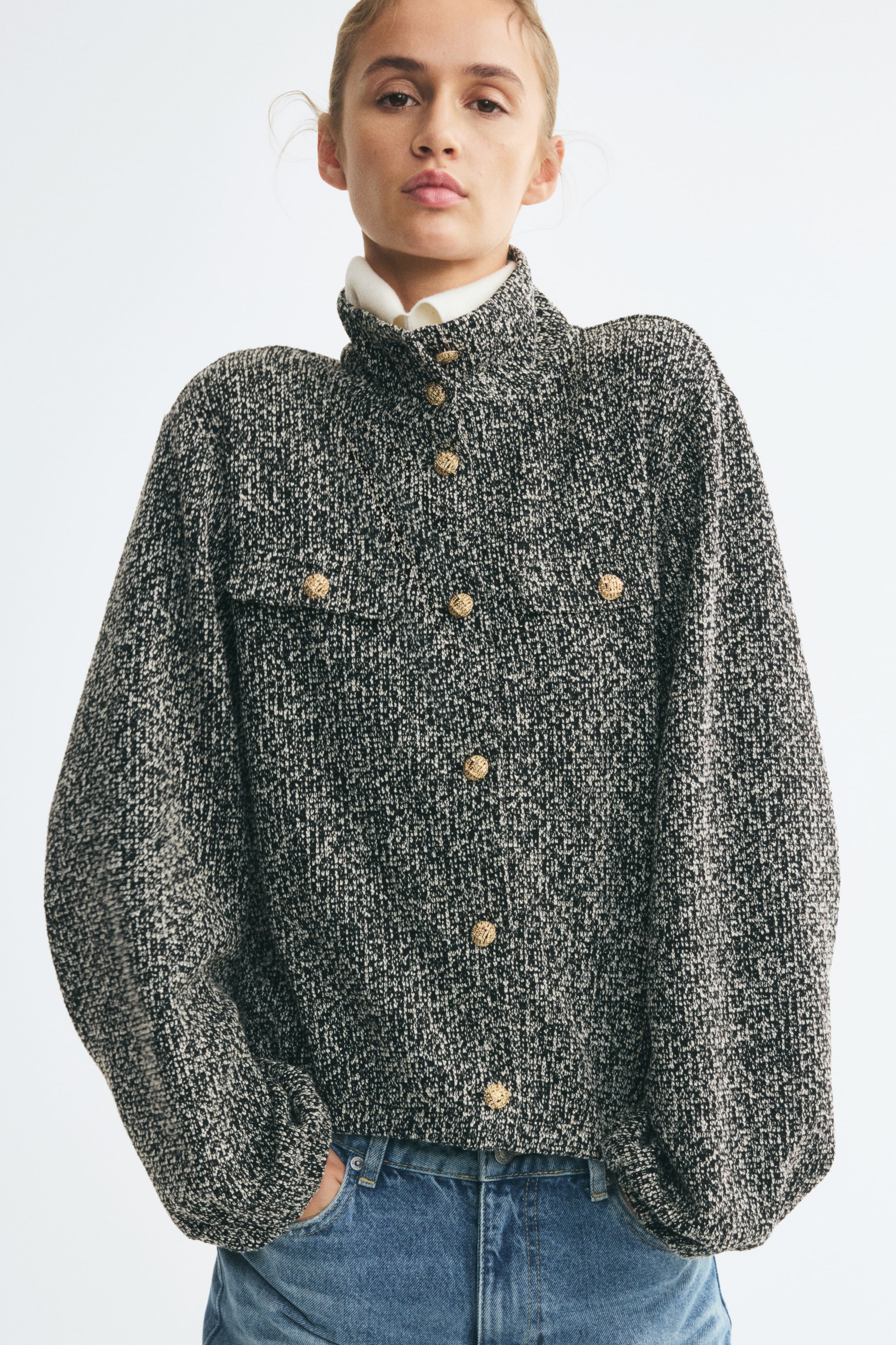 View larger image: The model is wearing a black and white textured tweed jacket featuring a high stand collar, gold-tone button closures, and wide sleeves. The jacket is layered over a white high-neck shirt and styled with blue denim jeans.