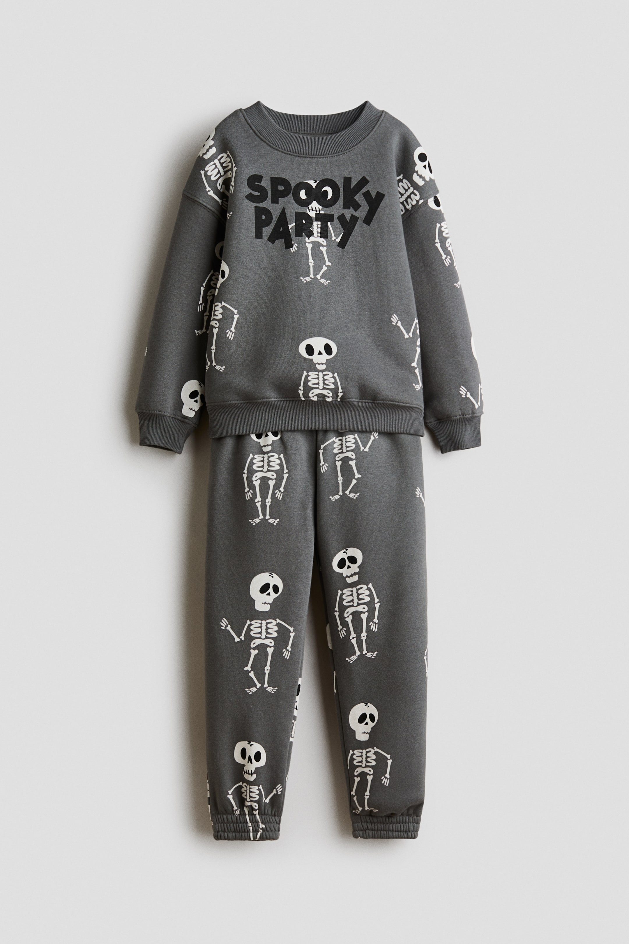 Grotere afbeelding bekijken: Een donkergrijze fleece sweatshirt en bijpassende trainingsbroekenset wordt frontaal getoond, met een all-over print van witte cartoon-skeletten. De sweatshirt met lange mouwen heeft een geribde ronde hals, verlaagde schoudernaden, geribde boorden en de woorden "SPOOKY PARTY" in zwart op de borst, terwijl de bijpassende broek elastische boorden aan de enkels heeft.