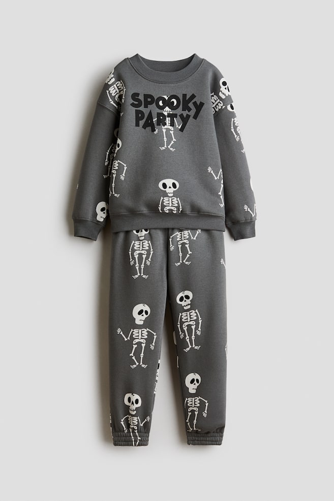 A dark grey fleece sweatshirt and matching track pants set is shown from the front, featuring an all-over print of white cartoon skeletons. The long-sleeve sweatshirt has a ribbed round neckline, dropped shoulders, ribbed cuffs, and the words "SPOOKY PARTY" printed in black on the chest, while the matching pants feature elasticated cuffs at the ankles. - 1