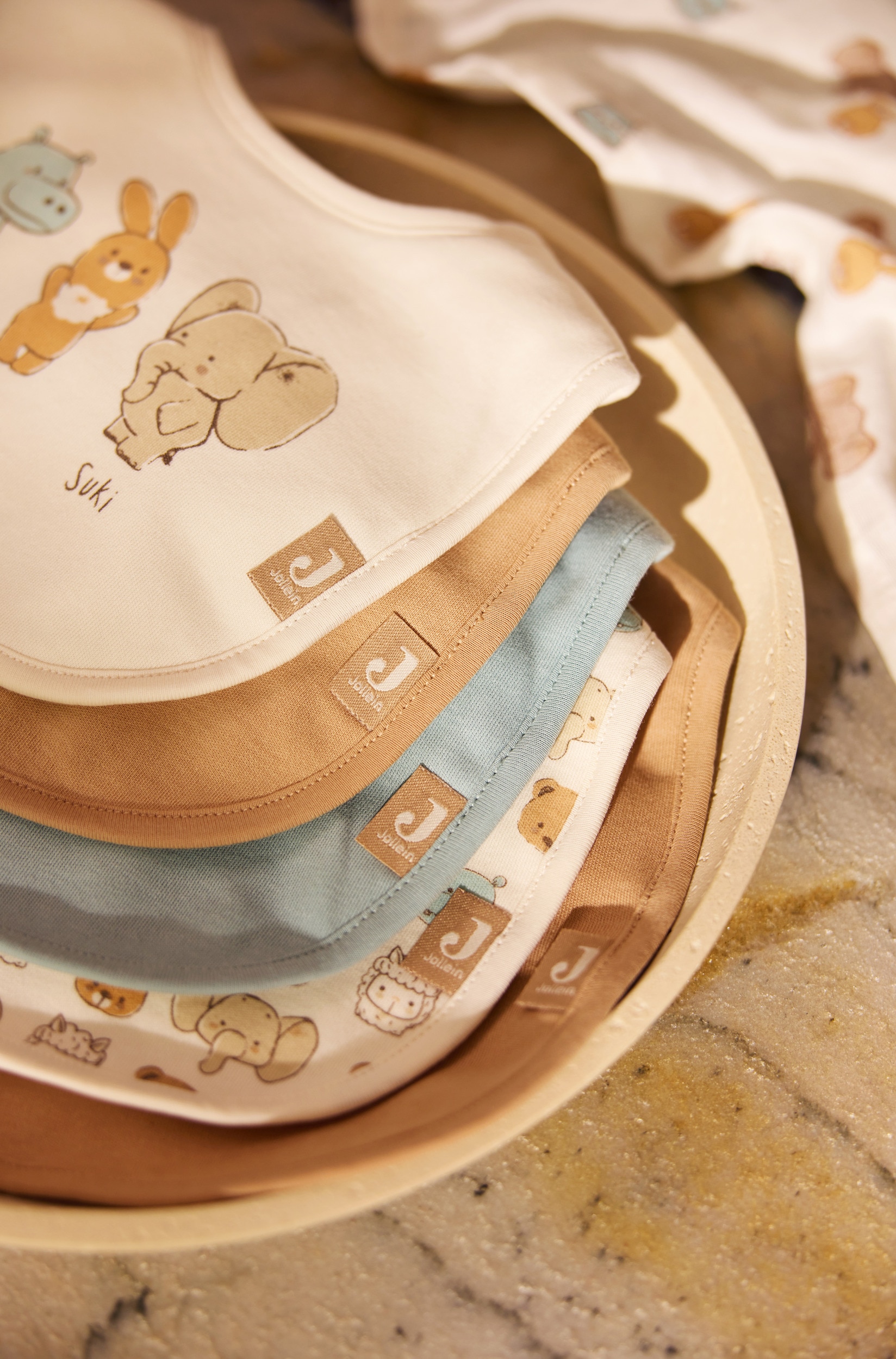 Een set van vijf slabbetjes voor baby's is diagonaal overlappend gerangschikt en toont een kleurenpalet van beige, gebroken wit en stoffig blauw. De selectie bestaat uit drie effen slabbetjes, Ã©Ã©n slabbetje met een all-over cartoon dierenprint en een beige slabbetje op de voorgrond met in het midden een alpaca-illustratie en daaronder de naam 'Paco'.