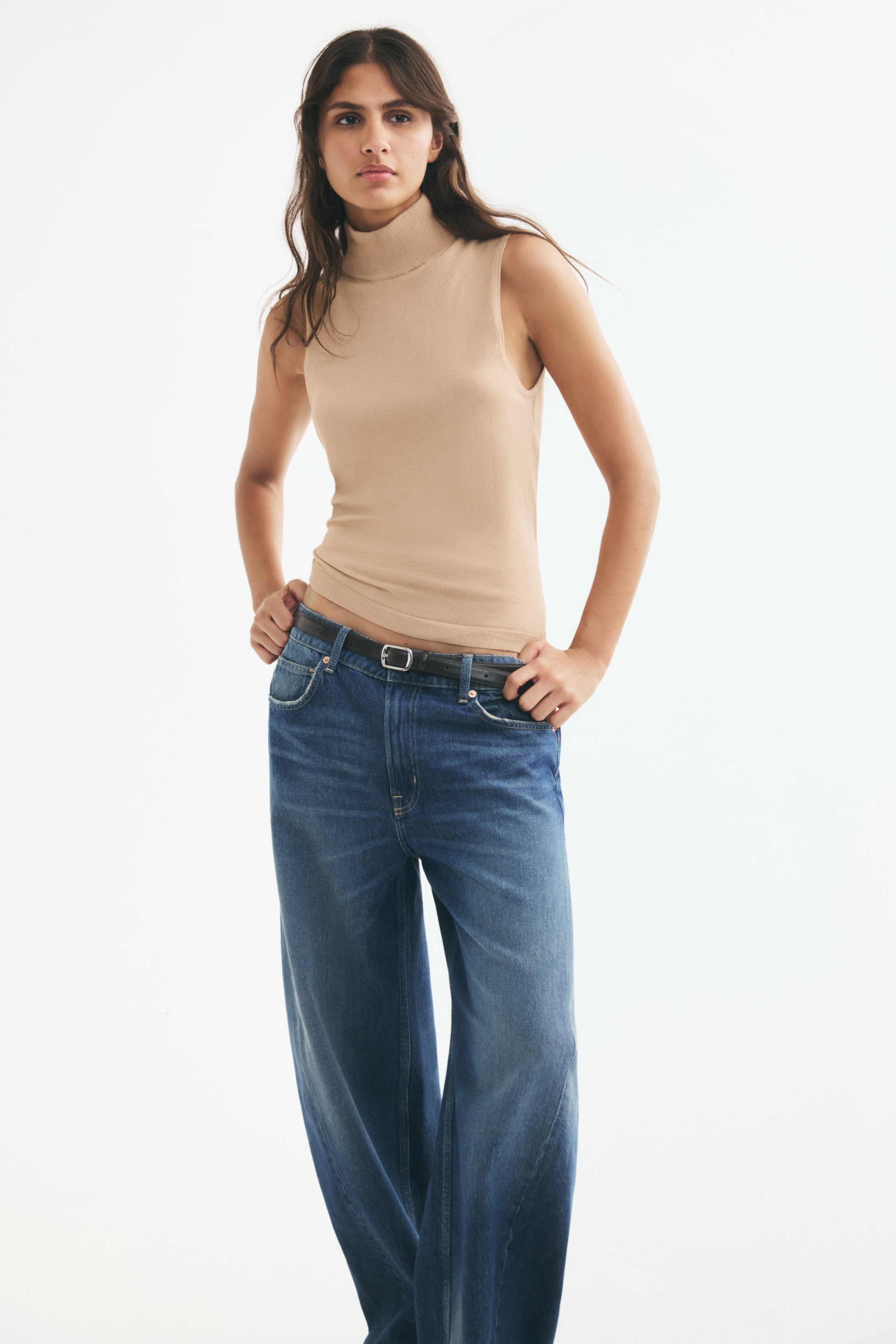 View larger image: A beige fitted knit vest with a ribbed turtleneck and ribbed hem is worn by the model. The vest is paired with blue wide-leg jeans and a black belt, with the model resting their hands near the waistband.