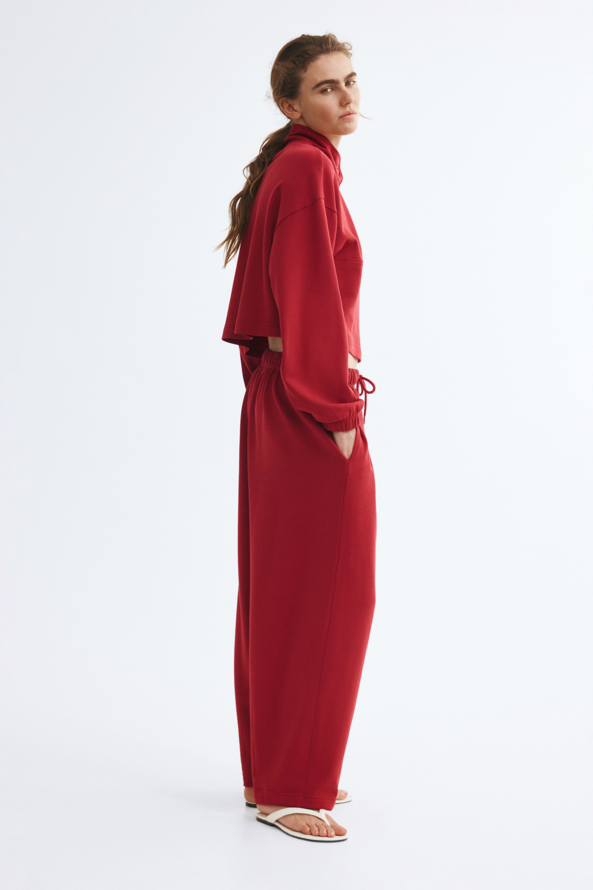 A pair of dark red trousers is shown from the front, featuring a gathered elasticated waistband with matching drawstrings and metal aglets. The trousers are shown in a relaxed, loose fit that tapers slightly towards the hem, which features subtle vertical seam detailing.