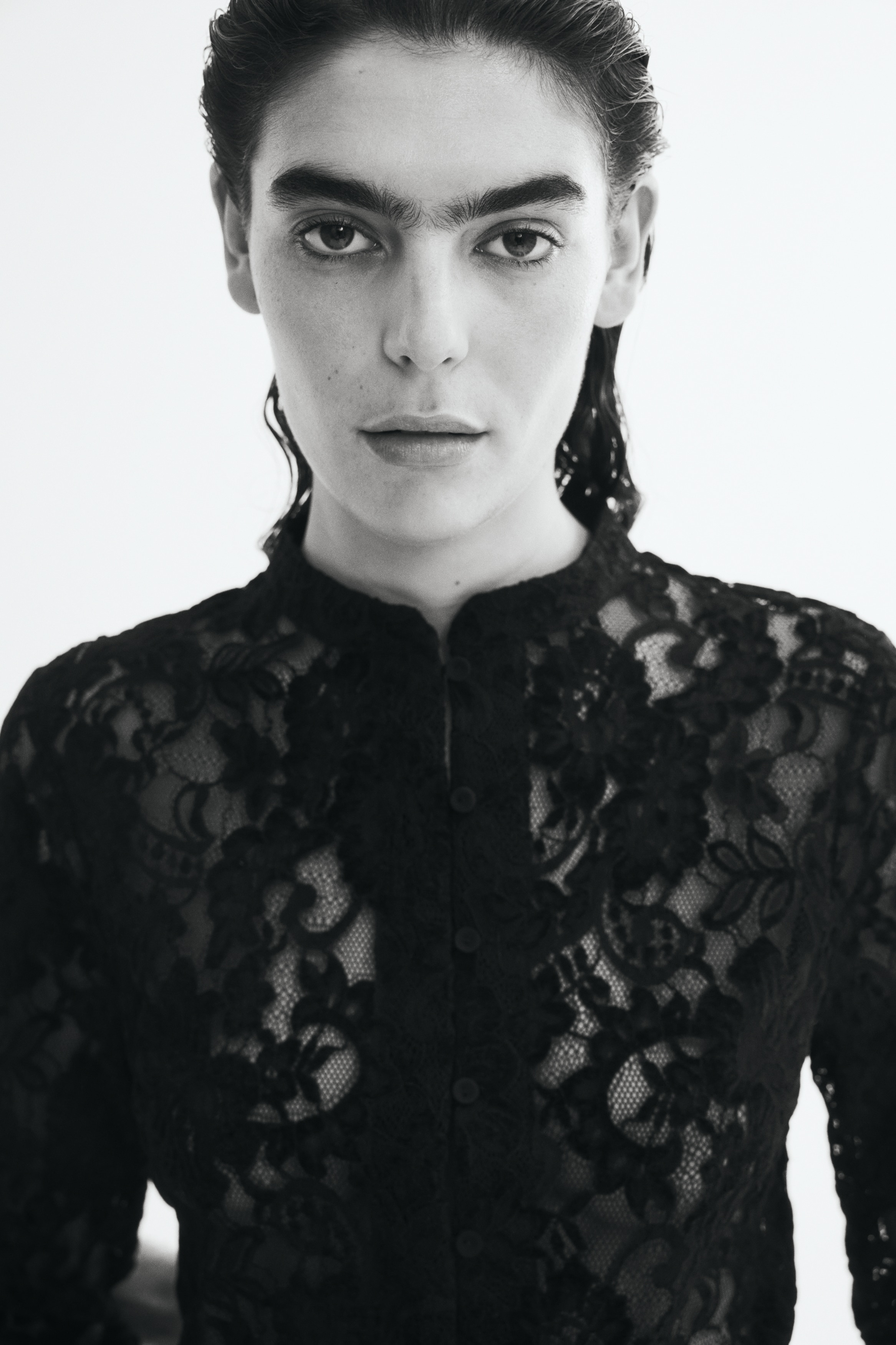 A black lace button-up top is shown from the front, featuring a high mock neck, long sleeves, and a straight, slightly cropped silhouette. The top features an intricate floral lace pattern throughout, complete with scalloped, eyelash-fringed edges along the hem and cuffs.