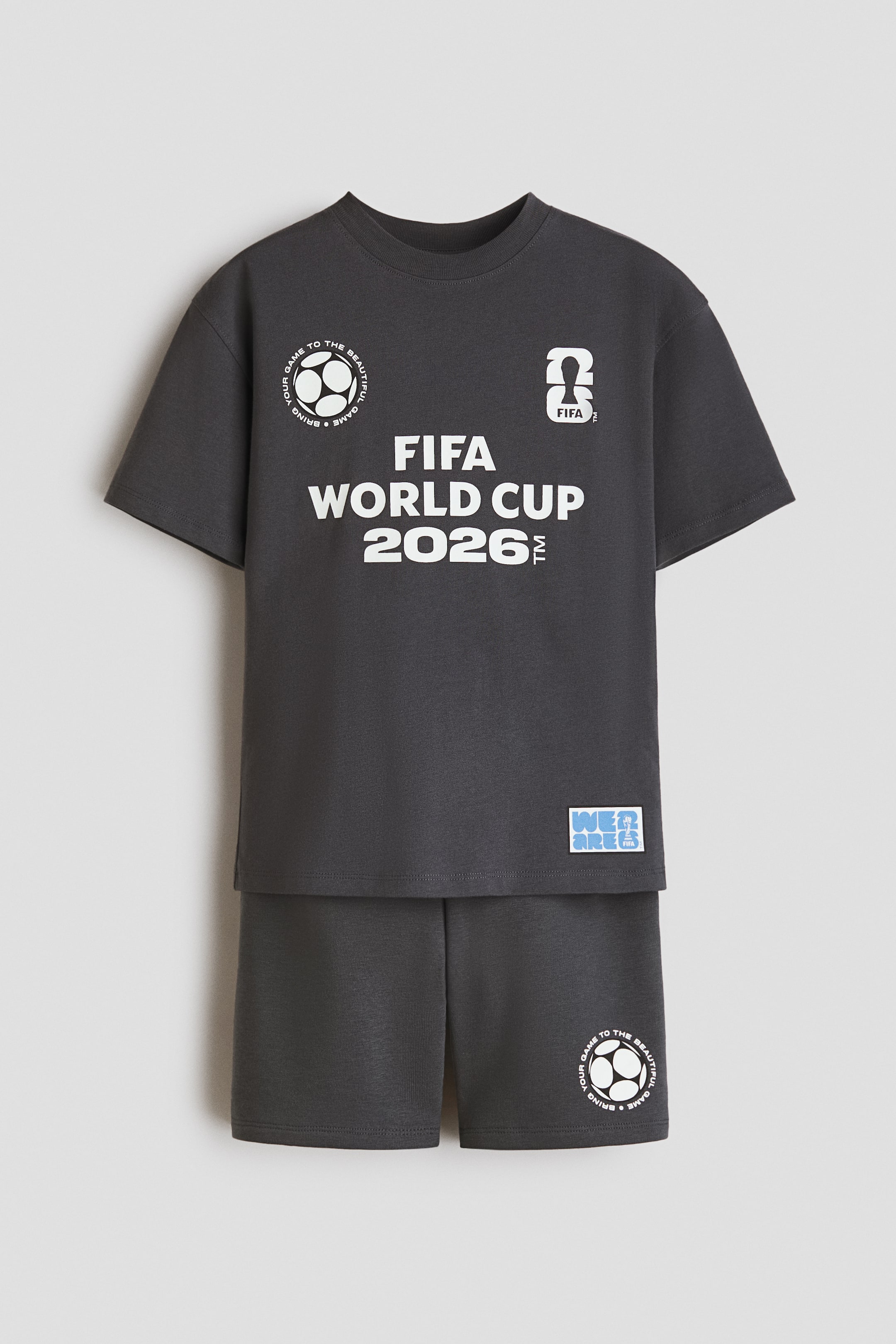 Visa större bild: Ett mörkgrått tvådelat set visas framifrån, bestående av en t-shirt med rund hals och korta ärmar samt matchande shorts. T-shirten har "FIFA WORLD CUP 2026" i vit text i mitten, en cirkulär fotbollsgrafik med text till vänster, och en "26 FIFA"-logotyp till höger, tillsammans med ett "WE ARE FIFA"-märke nära fållen. Shortsen har en cirkulär fotbollsgrafik med text på nedre högra benet.
