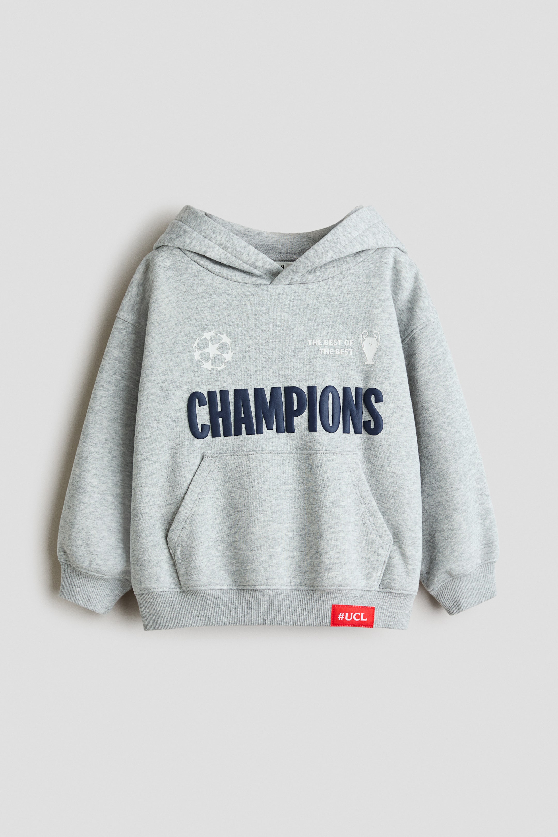 Visa större bild: En ljusgrå melerad hoodie visas framifrån med en omlottlagd huva, sänkta axlar och en känguruficka. Bröstet pryds av ett stort marinblått ”CHAMPIONS”-tryck tillsammans med vita fotbollsrelaterade grafiska detaljer, medan den ribbstickade nederkanten är accentuerad med en liten röd ”#UCL”-etikett.