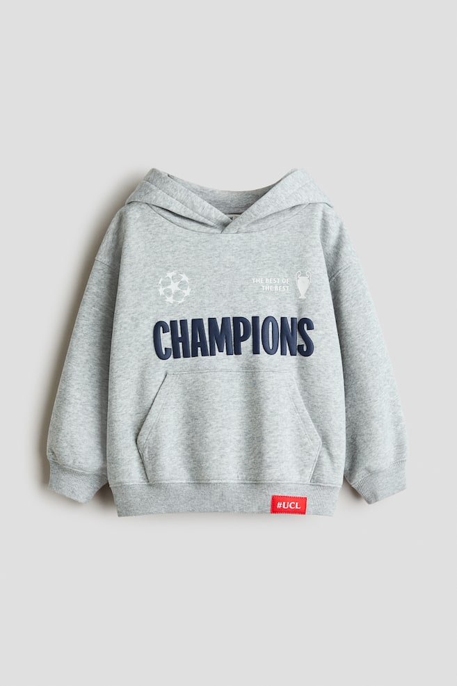A light grey mÃ©lange hoodie is shown from the front, featuring a wrap-front hood, dropped shoulders and a kangaroo pocket. The chest features a large navy blue "CHAMPIONS" print alongside white football-themed graphics, while the ribbed hem is accented with a small red "#UCL" tag. - 1