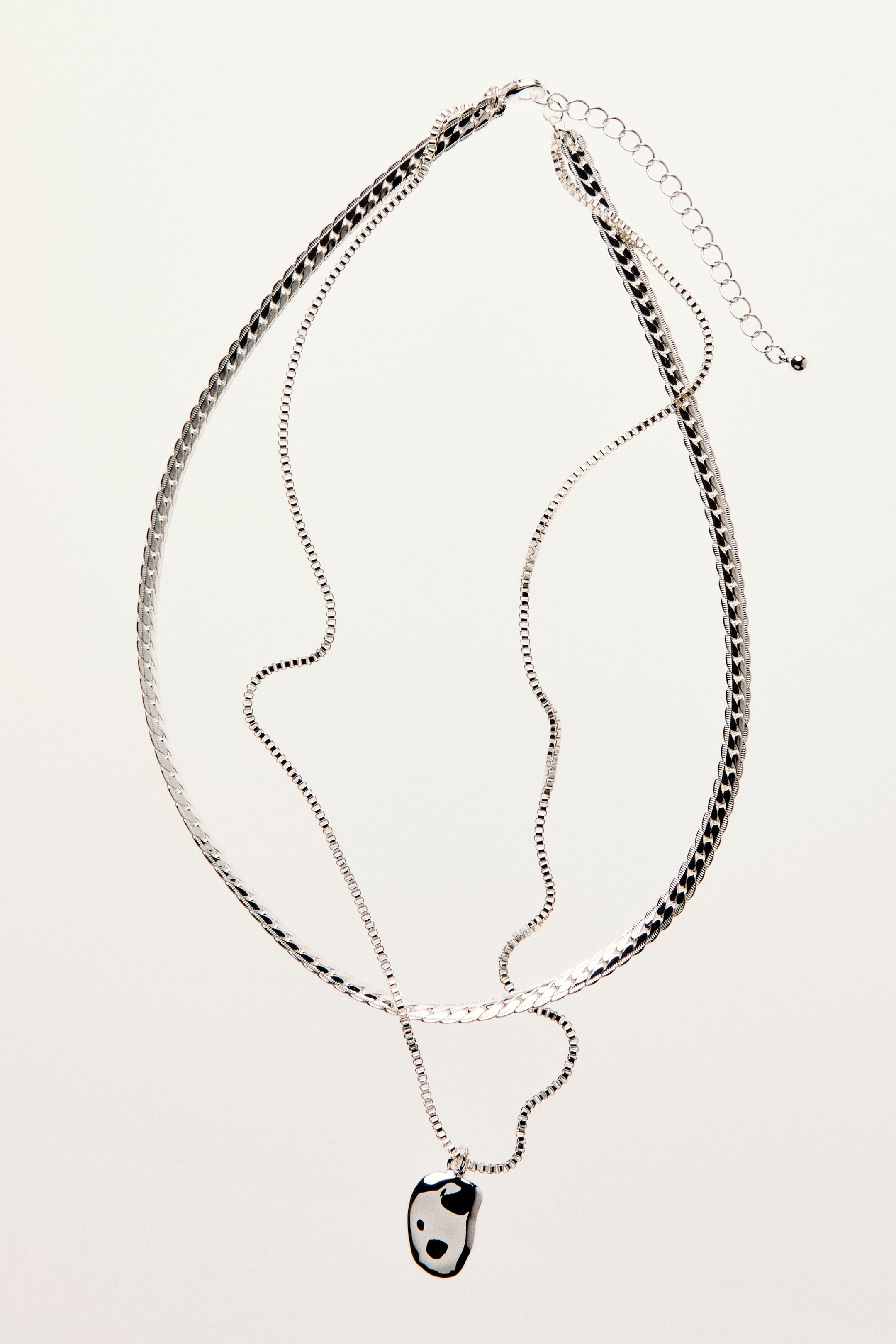 View larger image: A layered necklace showcasing two silver-toned metal chains, one with a flat, wide design and the other a slender box chain suspending an asymmetric, organically-shaped pendant with a black and white spotted pattern. The necklace is fitted with an adjustable extender chain and a trigger clasp.