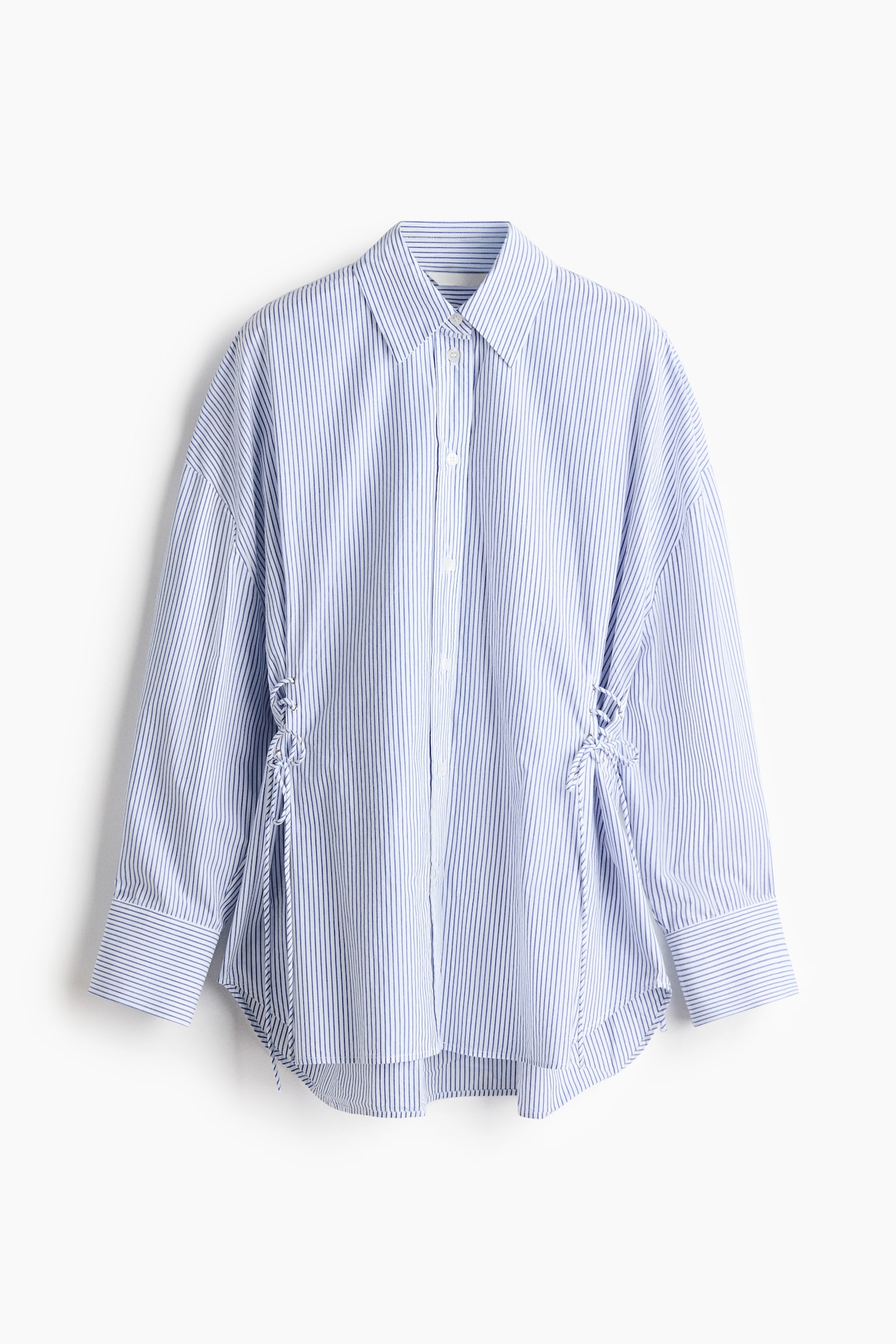 A blue and white vertically striped button-up shirt is hung facing front, showcasing a classic point collar, dropped shoulders, and wide cuffs. The shirt features adjustable lace-up detailing with tied cords on both sides of the waist and a curved high-low hem.