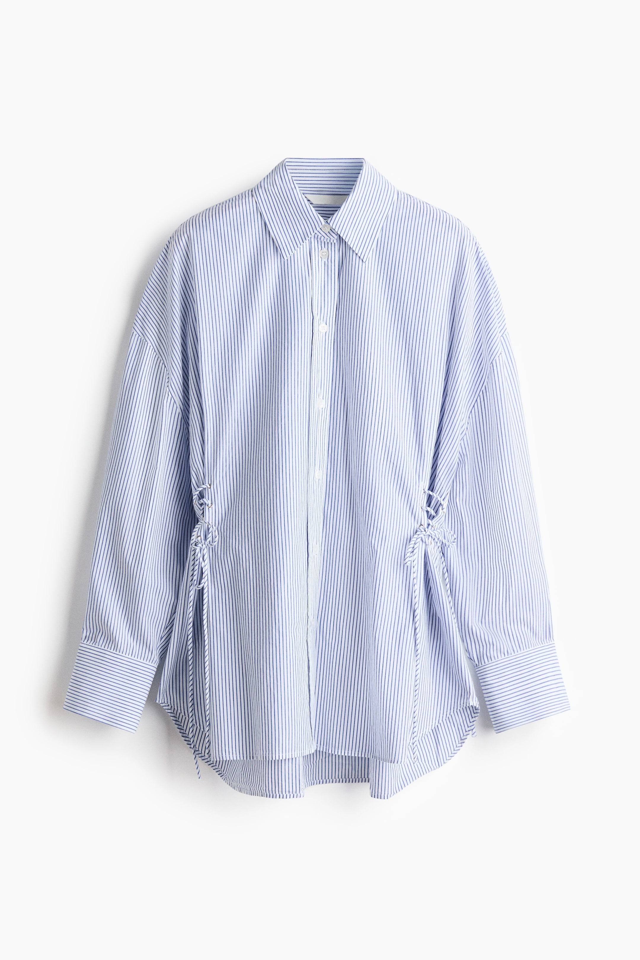 View larger image: A blue and white vertically striped button-up shirt is hung facing front, showcasing a classic point collar, dropped shoulders, and wide cuffs. The shirt features adjustable lace-up detailing with tied cords on both sides of the waist and a curved high-low hem.