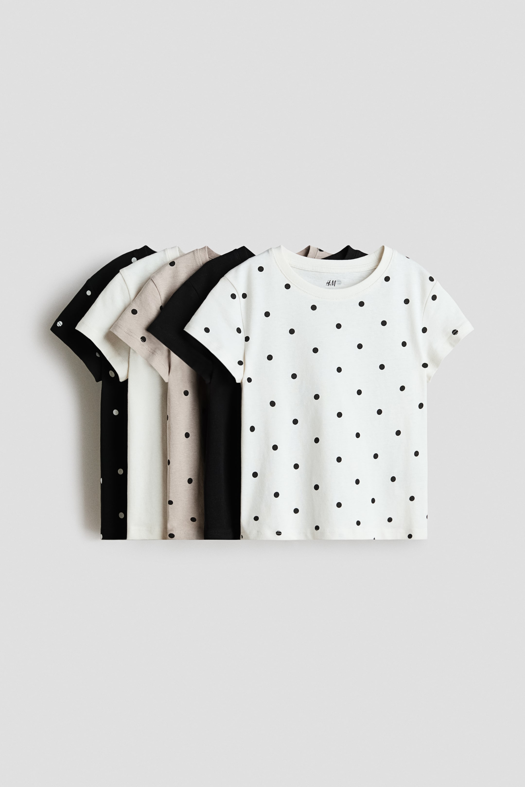 View larger image: T-shirts crafted from soft cotton jersey, featuring round, ribbed necklines and subtly dropped shoulders, are displayed front-facing. This set of five showcases solid hues in black, off-white, and beige. The foremost t-shirt stands out with an off-white base adorned with an all-over black polka dot design.