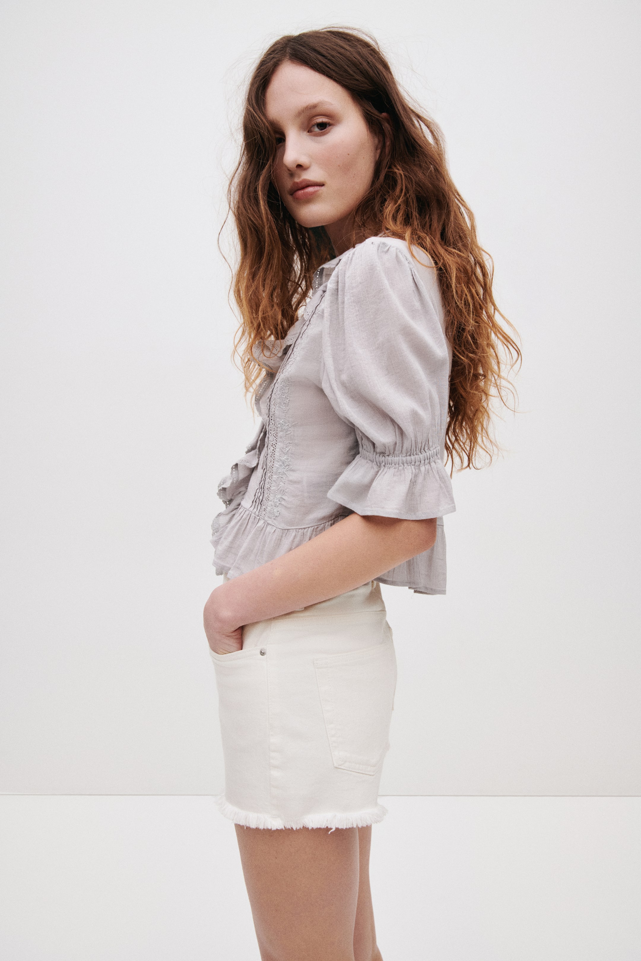 Ampliar la imagen: Una blusa gris claro se presenta desde un ángulo lateral leve, con mangas abullonadas y puños con volantes, alforzas, volantes a lo largo de la tapeta y detalles bordados. Se combina con shorts de mezclilla color crema que muestran un dobladillo deshilachado y bolsillos visibles al frente y atrás.