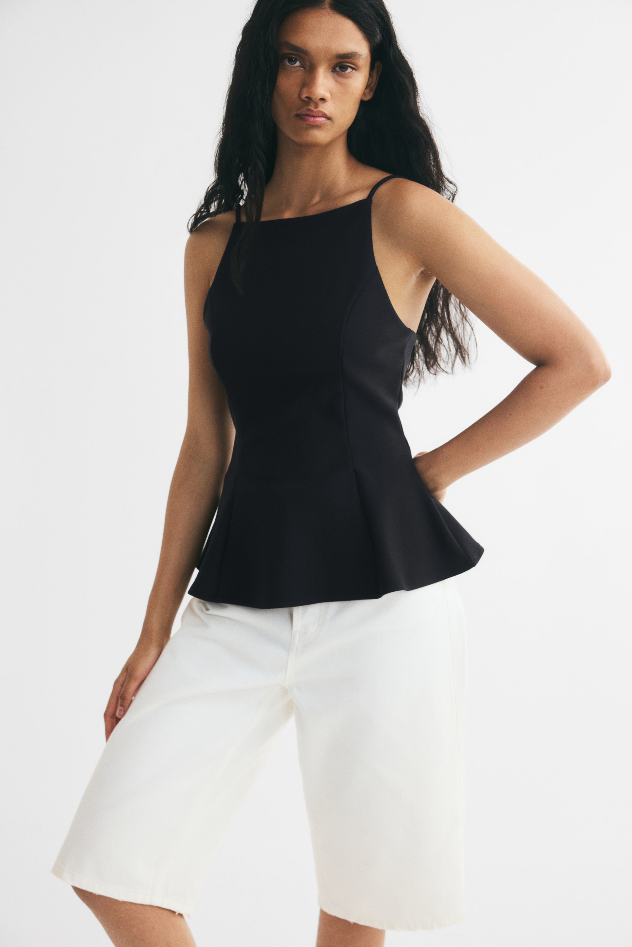 Ampliar la imagen: La modelo lleva un top peplum negro sin mangas, con tirantes delgados tipo espagueti, escote cuadrado, cuerpo entallado y dobladillo acampanado. El top se combina con pantalones blancos de pierna ancha.