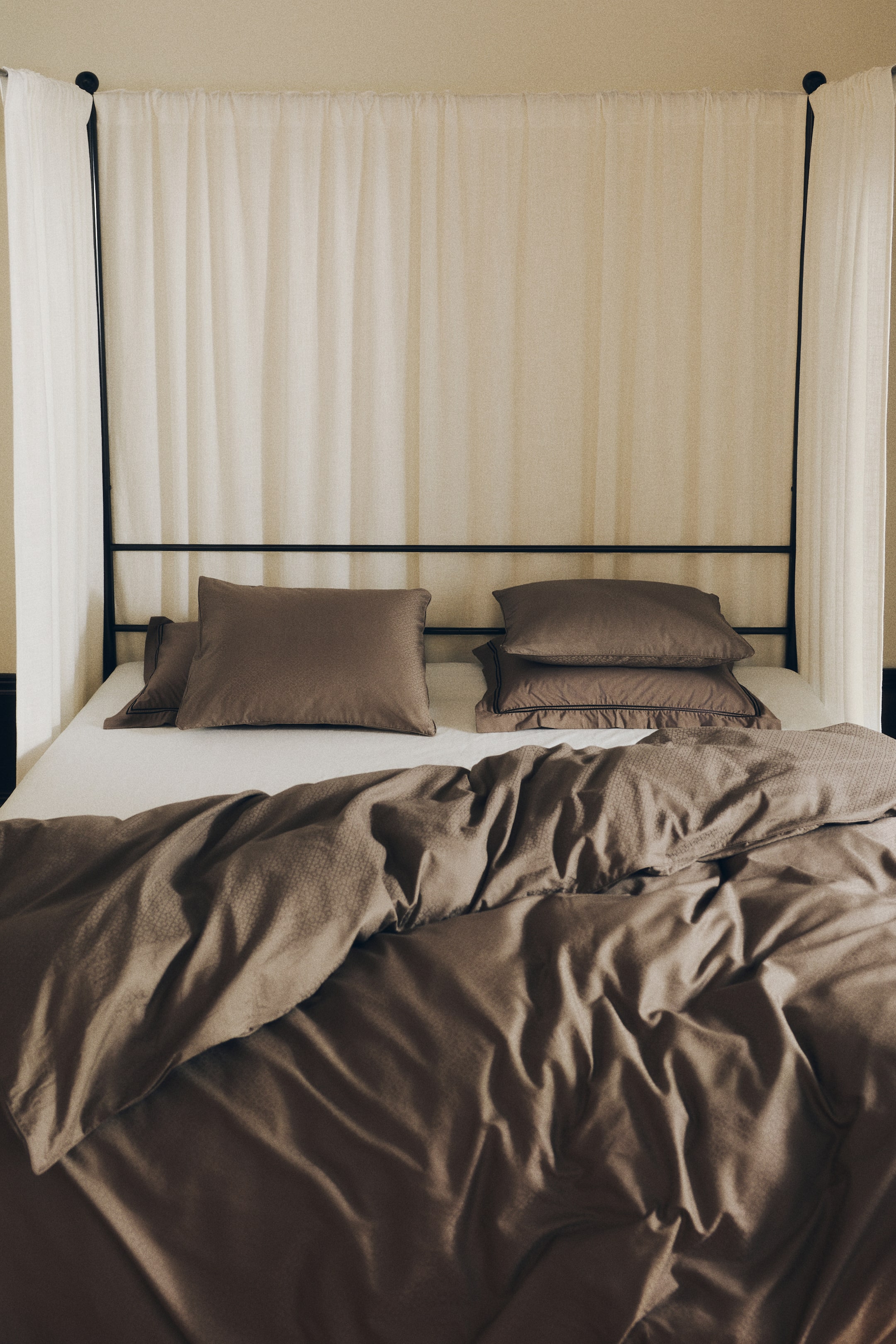 View larger image: A taupe cotton sateen duvet cover set is displaying a subtle sheen and a fine jacquard-woven geometric pattern on a bed. The set includes a loosely draped, rumpled duvet cover and matching pillowcases with thin dark piped edges arranged over a white sheet.