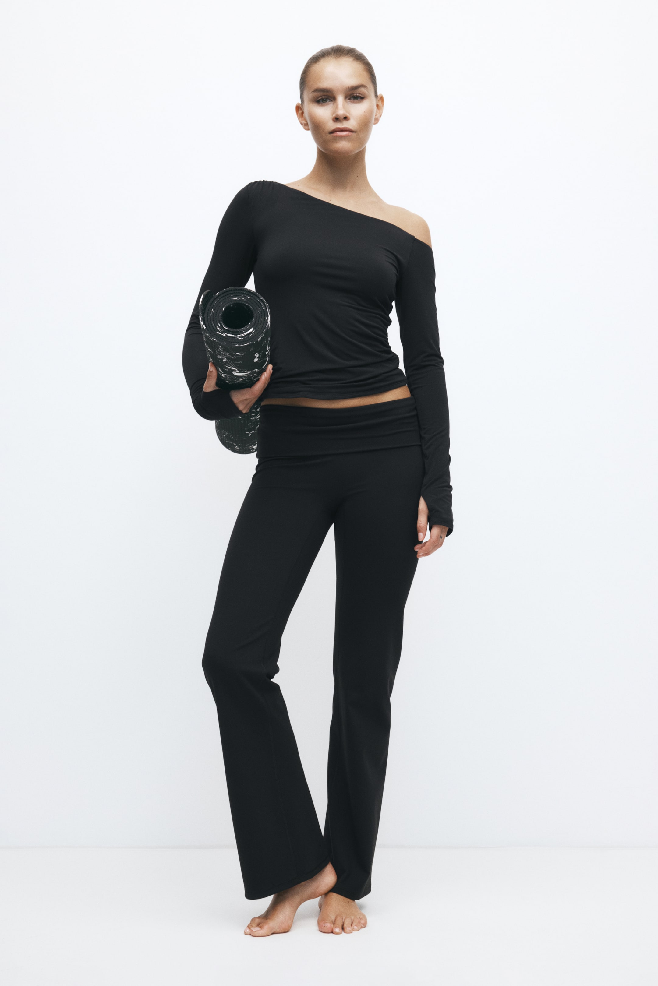 Büyük resmi görüntüle: Model, uzun kollu, omuzları açık bir üst ve katlanır bel bantlı, uyumlu ispanyol paça pantolondan oluşan siyah bir activewear seti giyiyor. Model, ayakta dururken bir kolunun altında rulo haline getirilmiş, siyah-beyaz desenli bir yoga matı tutuyor.