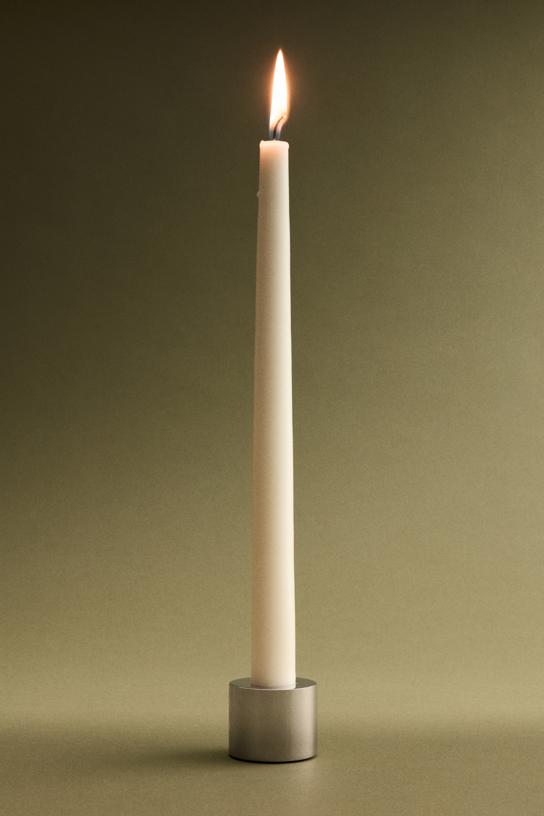 View larger image: An off-white taper candle stands upright, its top burning with a bright flame, and is placed in a cylindrical, brushed-silver candle holder.