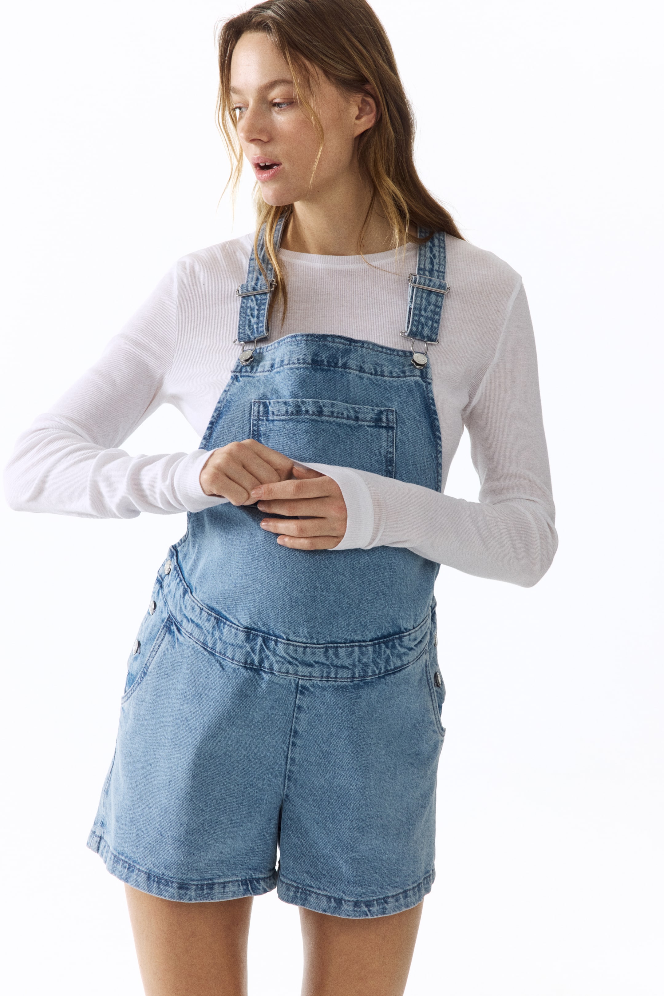 Größeres Bild ansehen: Hellblaue Denim-Shorts-Latzhosen mit verstellbaren Schulterträgern und silberfarbenen Beschlägen, einer Latzfront mit großer aufgesetzter Tasche und seitlichen Knopfverschlüssen an den Shorts, die auch zwei vordere Eingrifftaschen und zwei hintere aufgesetzte Taschen umfassen. Darunter wird ein weißes langärmliges Oberteil getragen.