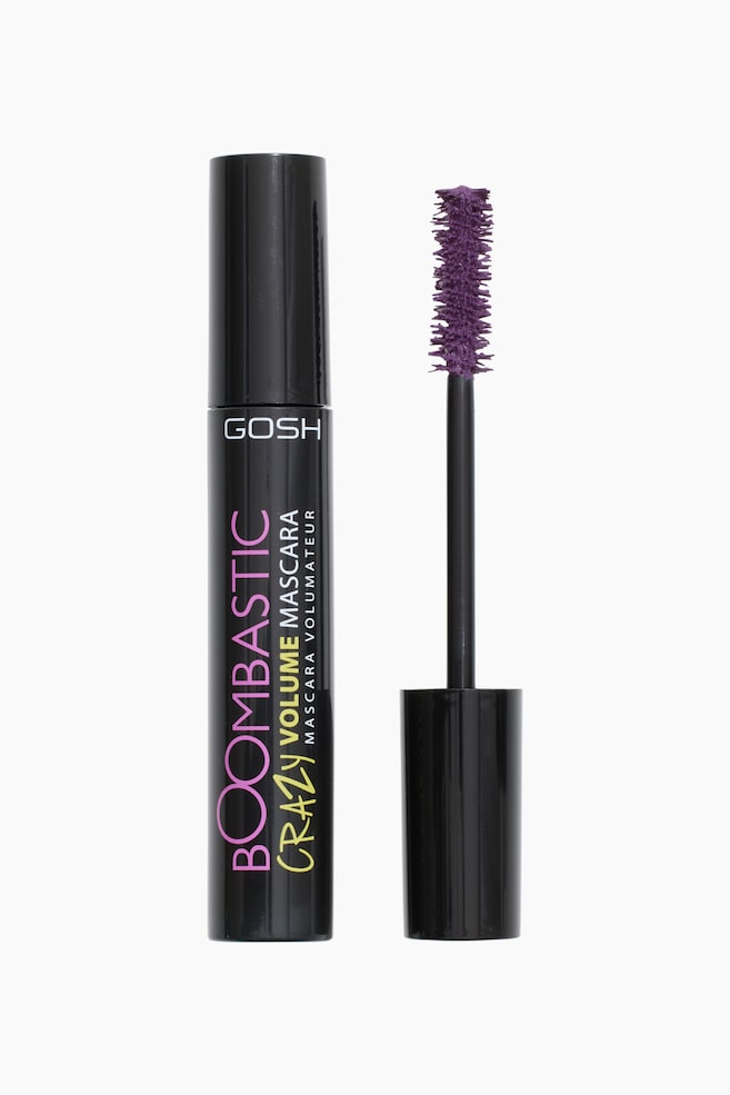 Et sort cylindrisk mascararør står oprejst, placeret til venstre for sin afmonterede børste. Røret har "GOSH" branding øverst og teksten "BOOMBASTIC CRAZY VOLUME MASCARA" vertikalt på siden. Børsten, med en sort stang, viser en tæt, spids lilla børste på sin spids. - 1