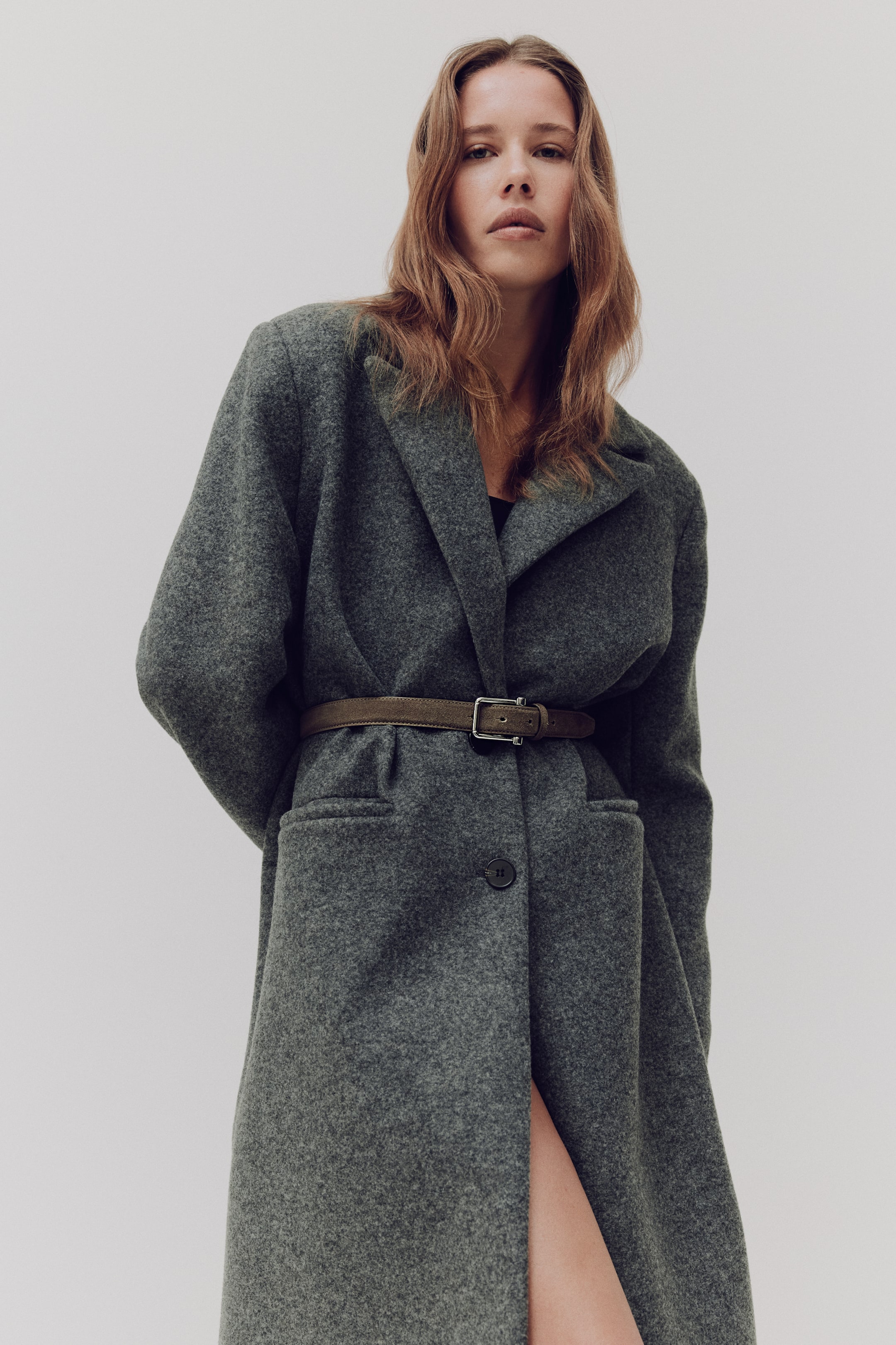 View larger image: The dark gray-green coat features wide notch lapels, two flap pockets, and a single visible button. A thin olive-green belt with a silver buckle cinches the coat at the waist, creating a fitted silhouette.