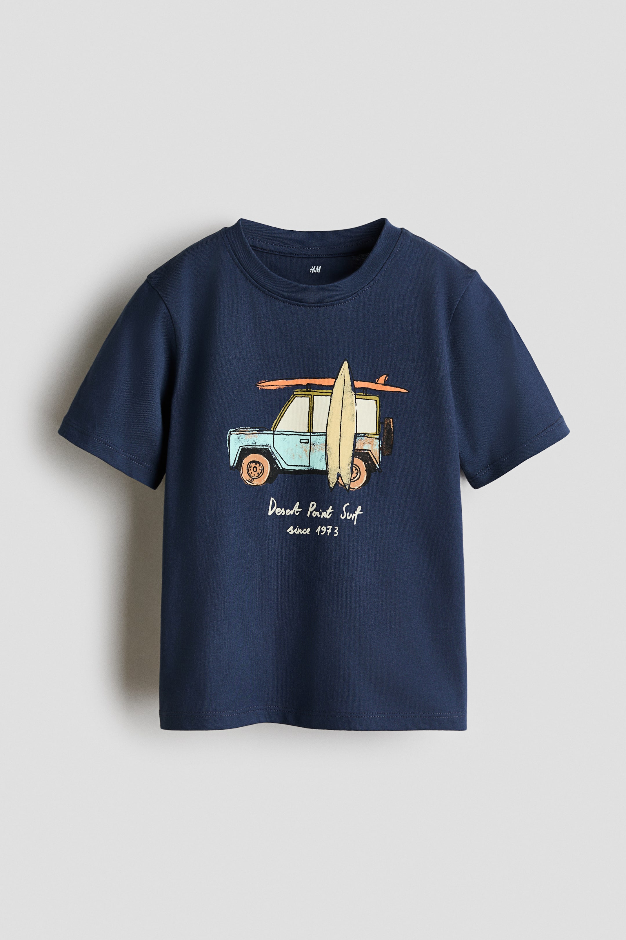 Grotere afbeelding bekijken: Een donkerblauw T-shirt in zachte katoenen jersey toont zijn voorkant. Het heeft ribbels rond de halslijn, verlaagde schouders en een recht gesneden zoom. Een grafische print op de voorkant toont een lichtblauw off-road voertuig met surfplanken en de tekst 'Desert Point Surf since 1973'.