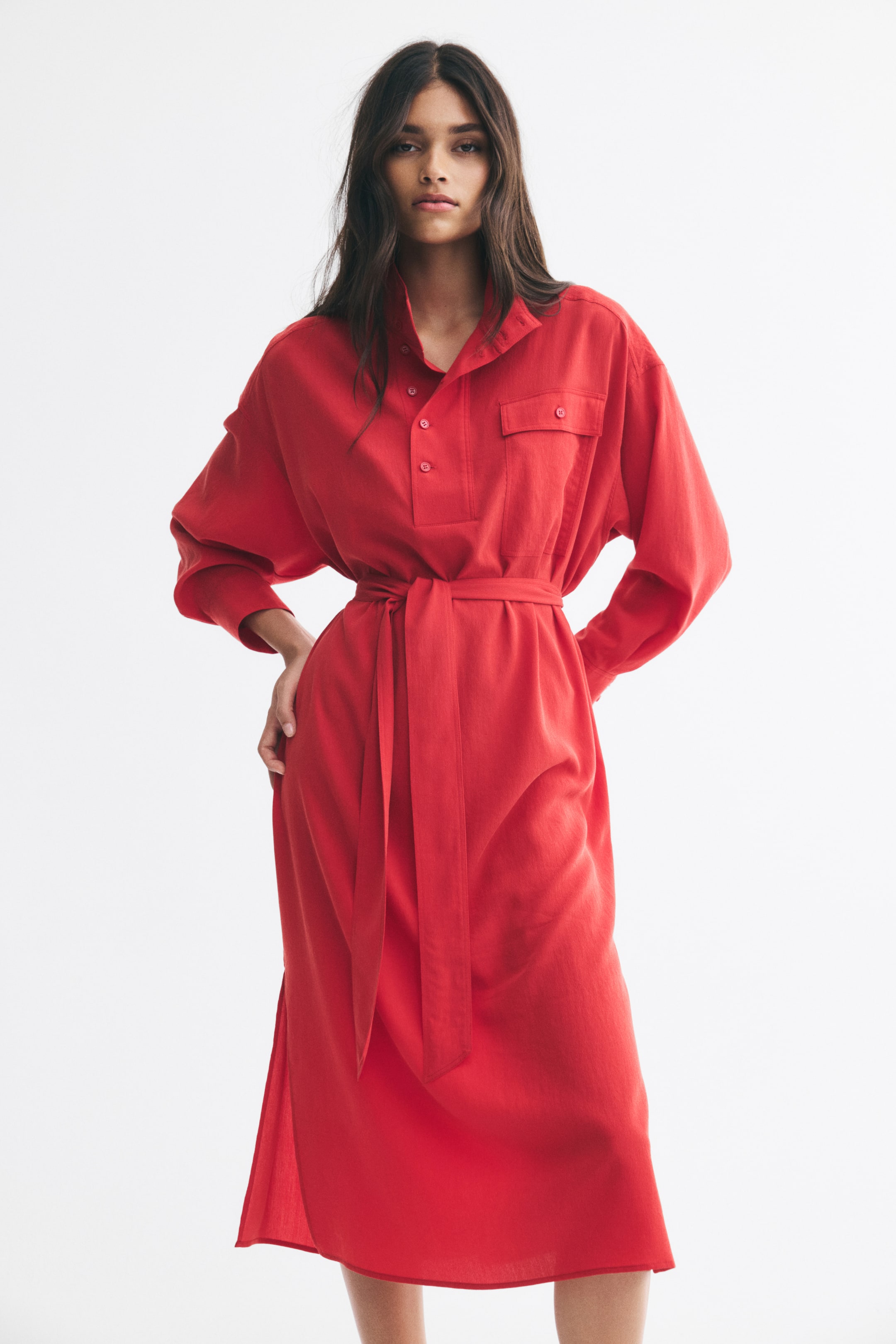 View larger image: The model is wearing a red long-sleeve tie-belt dress, featuring a high collar with a buttoned placket, a single chest pocket, and a matching fabric belt tied at the waist. The midi-length dress falls to the calves as the model stands with one hand resting on her hip.