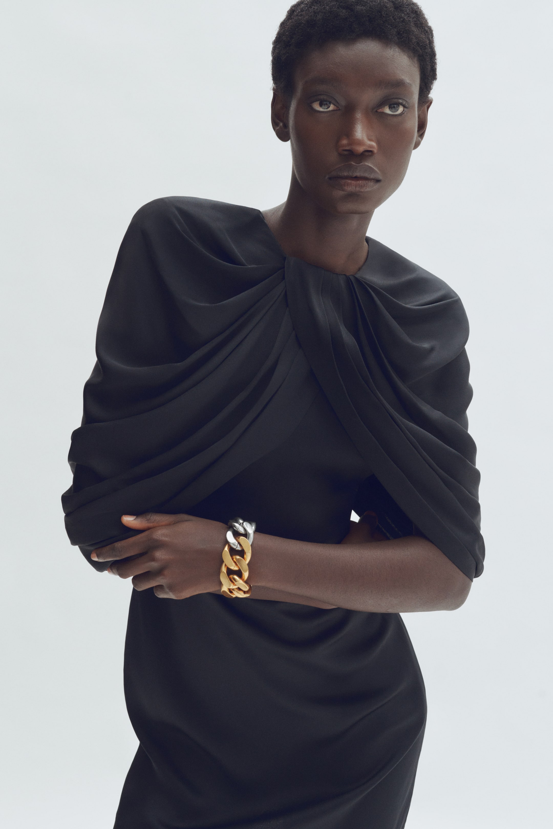 View larger image: A black garment showcases a draped high neckline with a central knot and voluminous sleeves, viewed from the front. A two-tone chain link bracelet in gold and silver is elegantly worn over the sleeve.