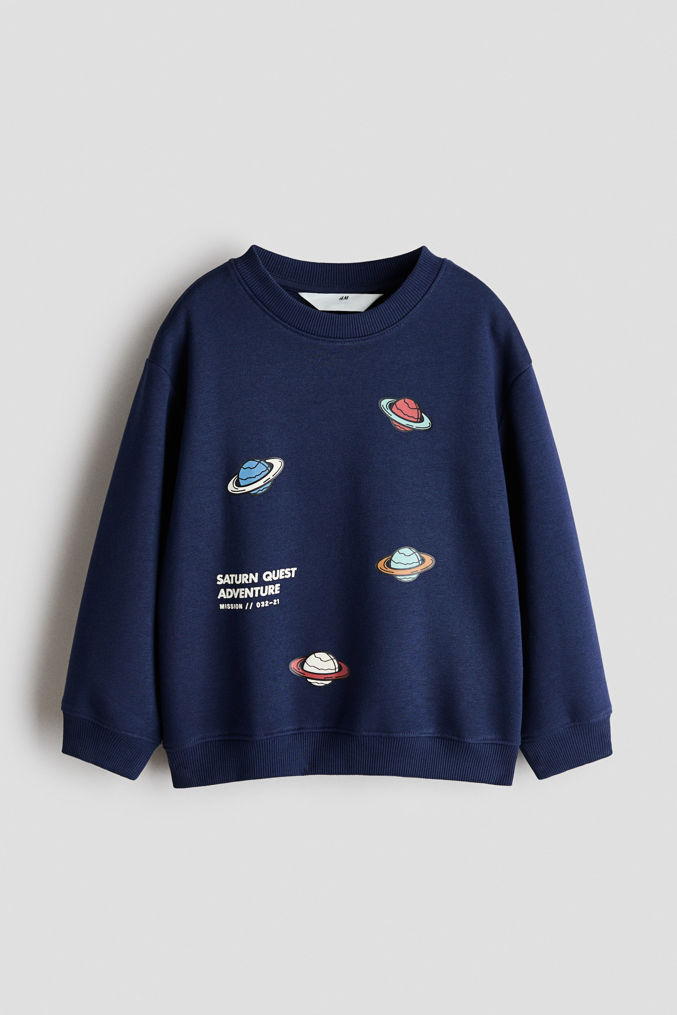 Xem ảnh lớn hơn: Một chiếc áo sweatshirt màu xanh navy được chụp từ phía trước, với cổ tròn bo gân, vai rơi và bo tay áo cùng gấu áo bo gân. Mặt trước nổi bật với họa tiết in gồm bốn hành tinh nhỏ nhiều màu có vòng và dòng chữ trắng "SATURN QUEST ADVENTURE MISSION // 032-21".