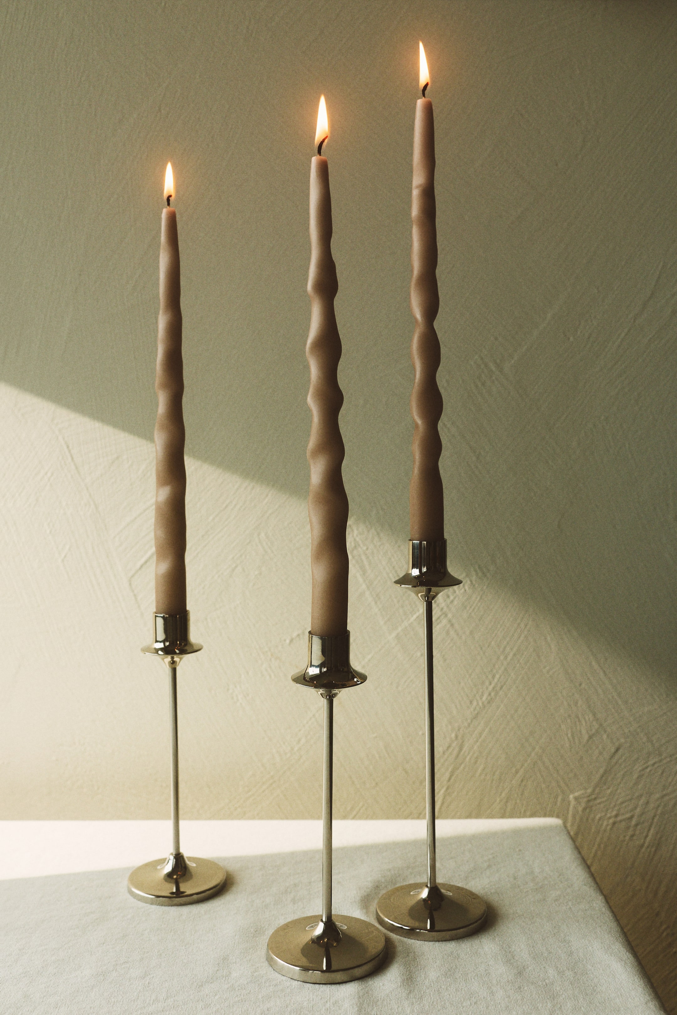 View larger image: Three lit taupe candles are standing upright in polished metallic candle holders, facing forward. Each candle displays a wavy, irregular texture along its length, while the holders feature slender stems and round bases.