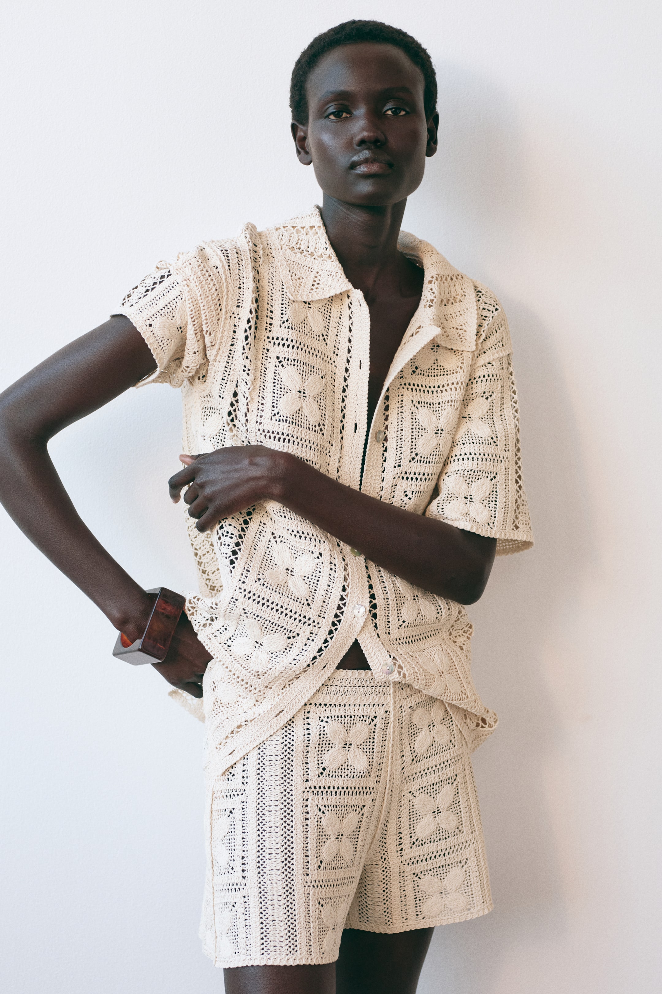 View larger image: A cream crochet two-piece set is being worn, consisting of a short-sleeve collared shirt and matching shorts. The shirt features a button-front, and both pieces display a repeating openwork square motif pattern with a central floral design.
