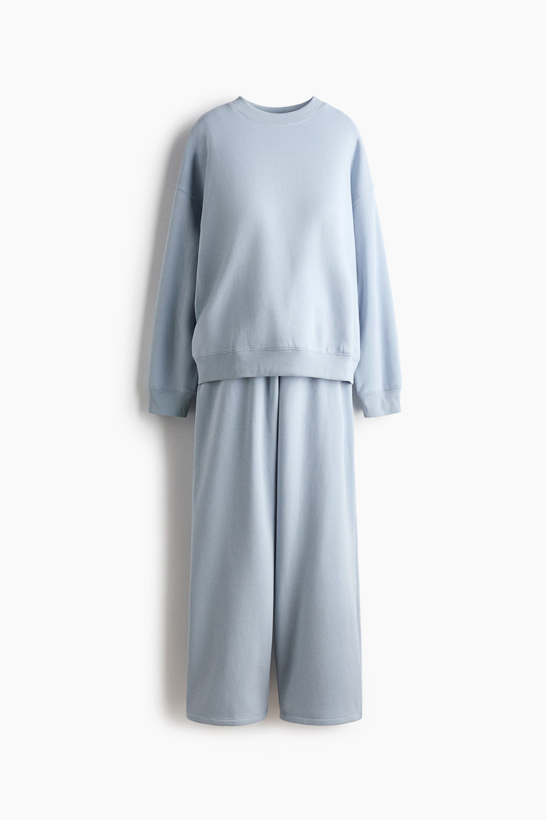 Grotere afbeelding bekijken: Een lichtblauwe loungewearset is van voren weergegeven, bestaande uit een sweatshirt met ronde hals en relaxte pasvorm en een bijpassende broek met wijde pijpen. Het sweatshirt heeft verlaagde schoudernaden, geribde boorden en een geribde zoom, terwijl de broek los en recht valt.