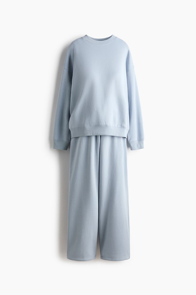 Ett ljusblÃ¥tt loungewear-set visas framifrÃ¥n och bestÃ¥r av en avslappnad sweatshirt med rund hals och matchande vida byxor. Sweatshirten har sÃ¤nkta axlar, ribbade muddar och en ribbad nederkant, medan byxorna faller i en lÃ¶s, rak silhuett. - 1